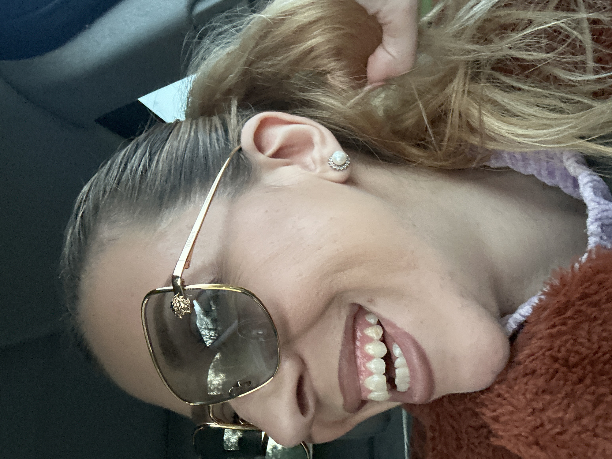 A woman is smiling, with her head tilted. She is wearing gold-framed sunglasses with a visible Versace emblem. Her hair is styled in a bun. She has a pearl-like stud earring in her left ear. She is wearing a fuzzy brown garment. The image was taken indoors, possibly inside a car. No information exists to suggest that the image was taken in Basildon, United Kingdom.
