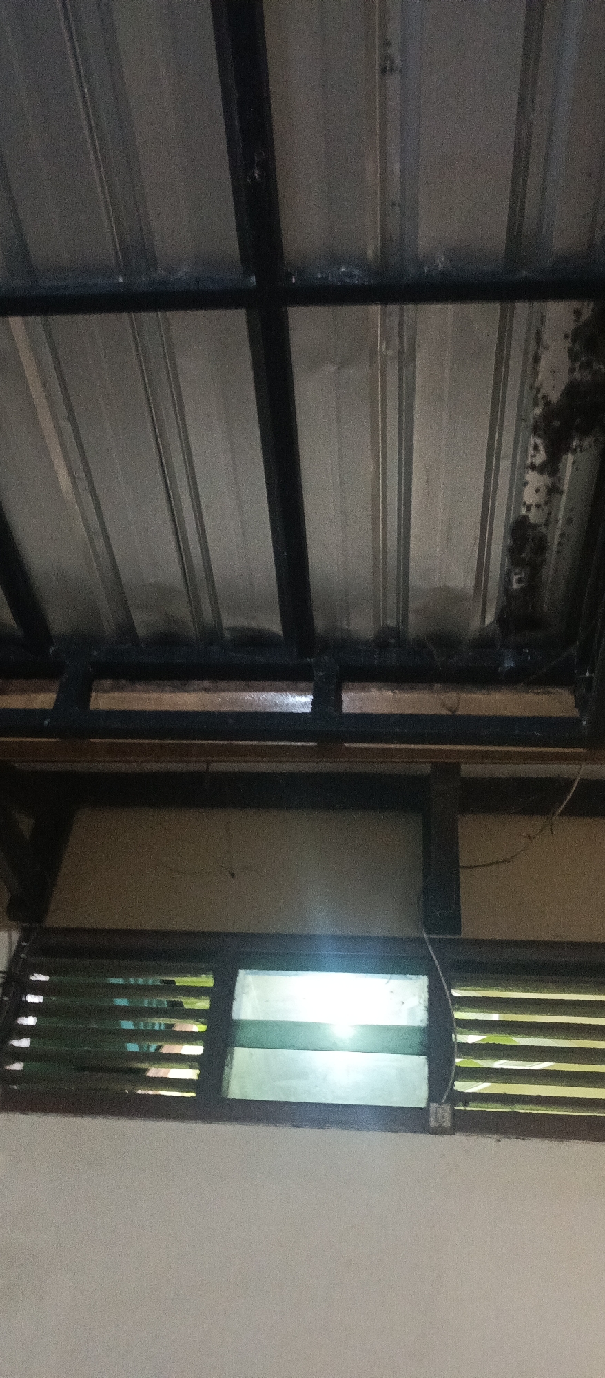 The image shows the underside of a metal roof with visible support beams. Below the roof, there is a window with horizontal slats, likely a louvered window. The window is divided into two sections, with the upper section appearing to be made of glass, and the lower section also featuring slats similar to the upper part. The wall below the window is light-colored and textured. The overall scene suggests an interior or sheltered exterior space, possibly part of a residential or commercial building in Kedung Dowo, Indonesia, given the context. There are no people or discernible actions depicted in the image. The lighting is dim, and there is some debris or discoloration on the metal roof structure.