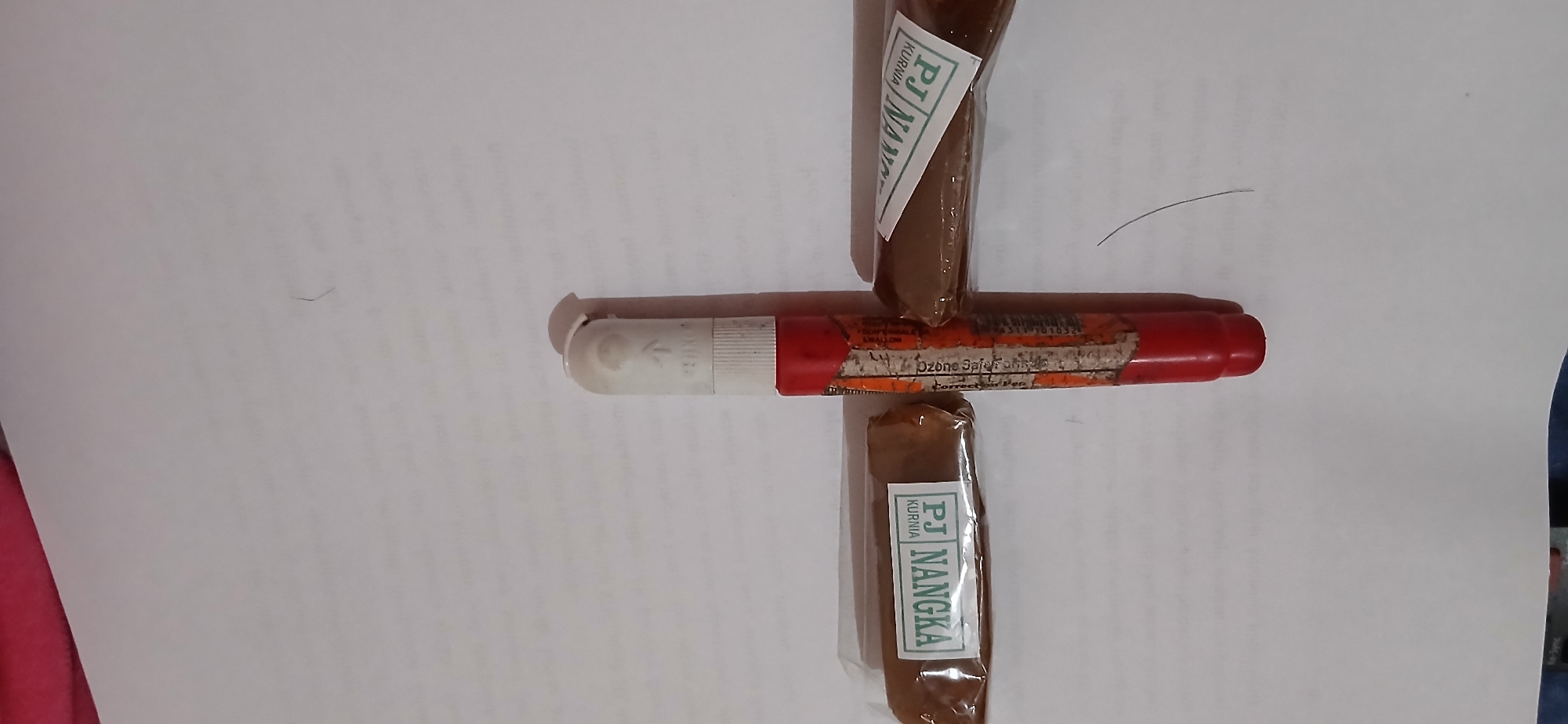 An overhead, eye-level shot shows two individually wrapped brown, rectangular objects on either side of a red and white marker. The marker is positioned vertically, with its white tip pointing up and its red base pointing down. The white tip is covered by a white cap. The marker has orange and yellow text printed on its side. The two brown objects appear to be food items, possibly snacks, and are wrapped in clear plastic. Each object has a white label with green text that reads "PJ NANGKA" and "KURNIA". A single dark hair is visible on the white surface below the marker. The background is a plain white surface, likely a table or paper, with some faint text visible in the distance. No people are present in the image. The scene is indoors, with the lighting appearing to come from above.
