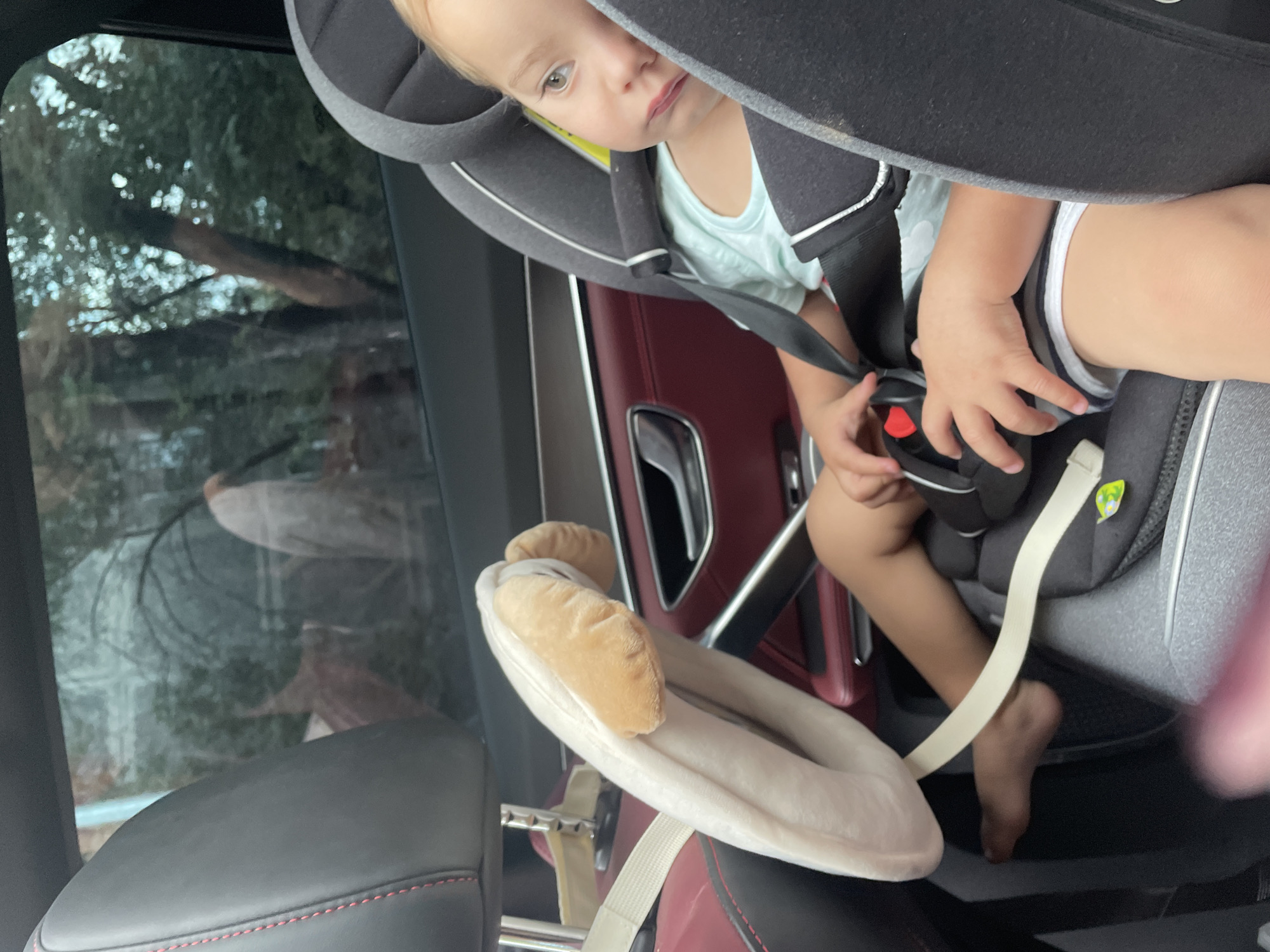 A young Caucasian child sits buckled in a car seat, looking to the right with a slightly pouty expression. The child is wearing a light blue t-shirt and striped shorts. A plush, toy-like accessory with tan ears is attached to the car seat in front of the child. The interior of the car is visible, with dark leather seats and a glimpse of trees through the tinted window, suggesting an outdoor setting in Almaty, Kazakhstan. The child's hands are near the car seat buckle. The lighting is natural, indicating daytime.