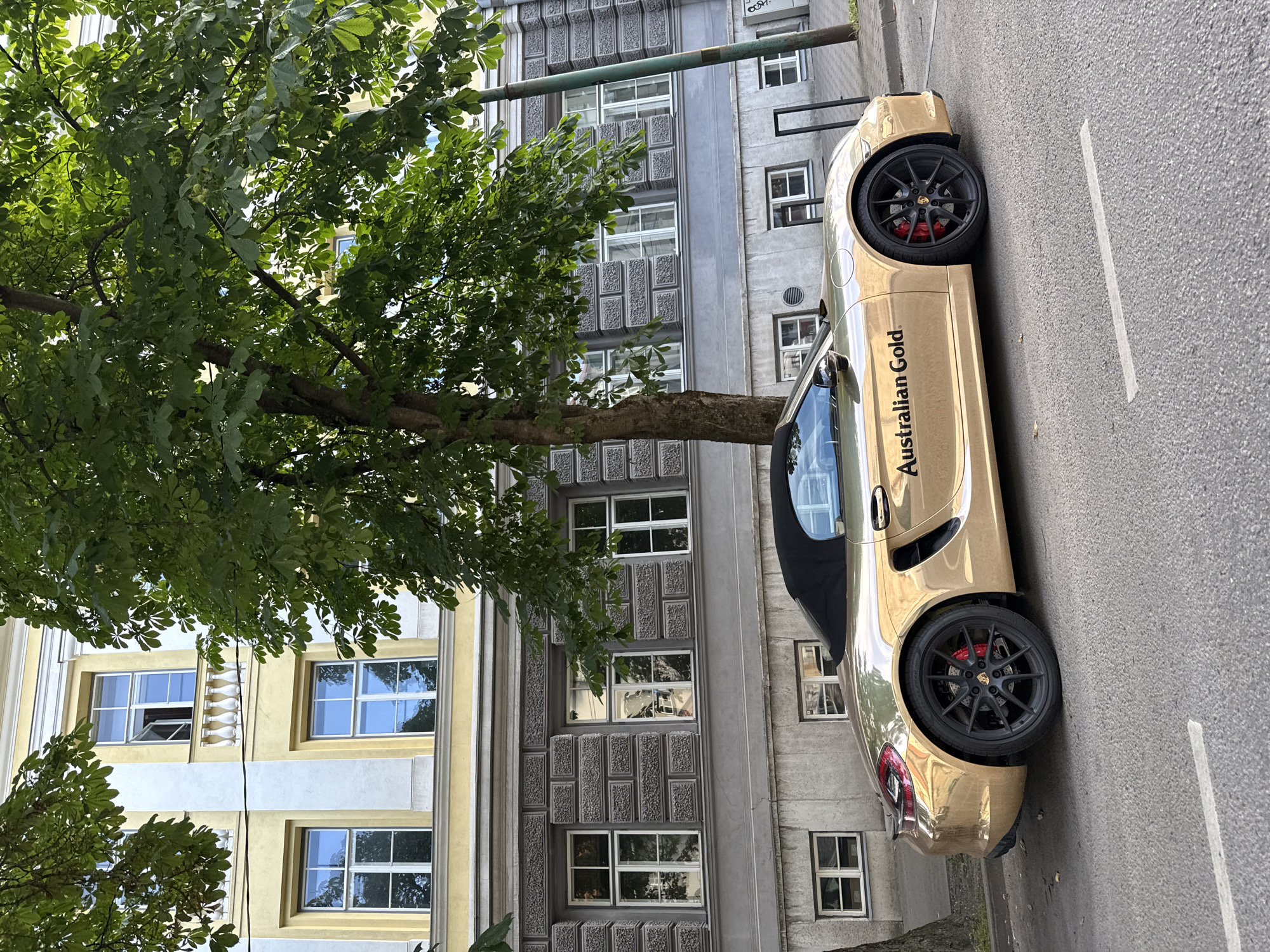 A gold Porsche Boxster convertible with a black soft top is parked on a city street. The car has black wheels with red brake calipers and the text "Australian Gold" is visible on the driver's side door. Behind the car is a multi-story building with yellow and gray facades, featuring numerous windows. A large tree with green foliage partially obscures the building and car. The street is paved with asphalt and a white line marks the center of the road. The sky is not visible in the frame.