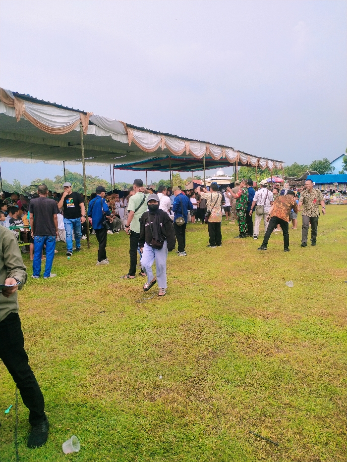 The scene takes place in Karangmalang, Indonesia, at the coordinates -7.47 latitude and 111.064 longitude. It features a large gathering of people under a series of white canopies with brown trim. The ground is covered in green grass. The people are casually dressed, some are standing around, others are talking, and a few are walking. There are a variety of objects in the scene including a few plastic chairs, a discarded plastic cup, and a small, abandoned black rope lying on the ground. The canopies are supported by metal poles. The sky is overcast. A few trees are visible in the background. A building is also visible in the background, though it's unclear what type of building it is.