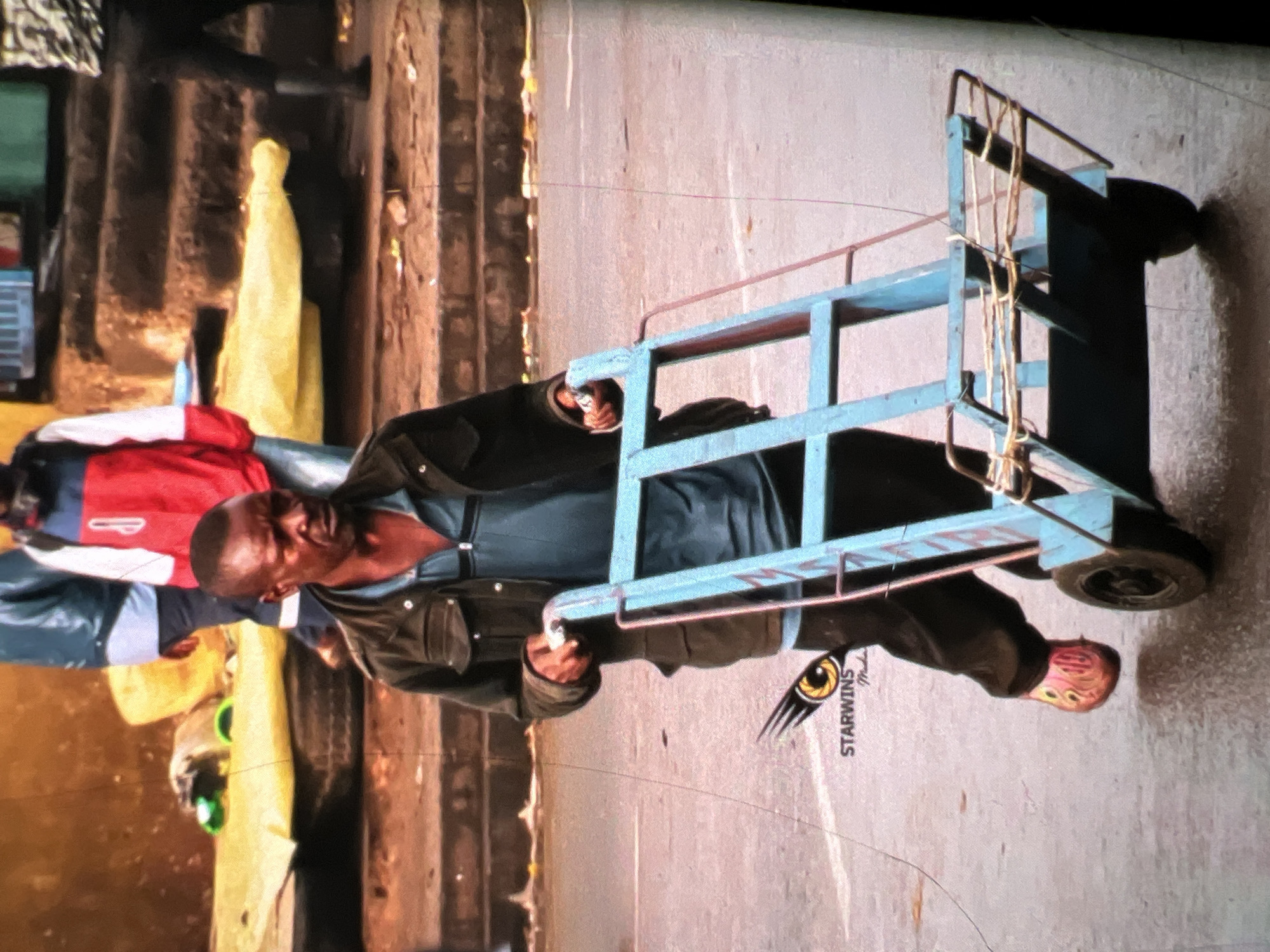 In Lagos, Nigeria, a man with dark skin and a short beard is pushing a blue hand truck. He is wearing a dark green jacket over a blue collared shirt and dark pants. He is also wearing red and yellow sandals on his feet. The hand truck is made of metal with a blue frame and a dark green platform. The background of the image shows a street scene with people in the distance. One person is wearing a red and white jacket, and another is wearing a black and white patterned shirt. There are also stairs and yellow tarps in the background. The ground appears to be wet, suggesting recent rain. The overall scene depicts a typical street environment in Lagos.