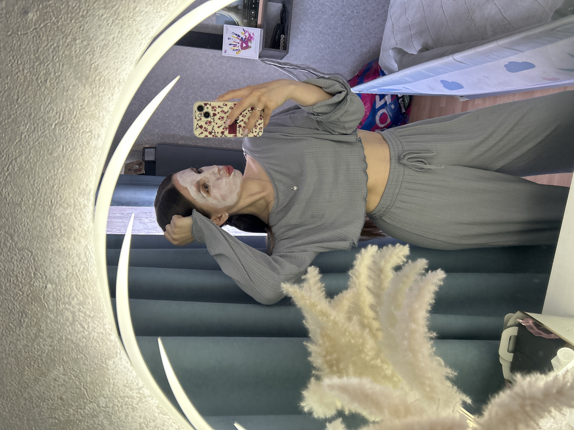 A woman is captured in a mirror selfie in an indoor setting in Klin, Russia. She is wearing a light greyish-green ribbed loungewear set, consisting of a long-sleeved crop top and matching wide-leg pants with a drawstring waist. A white face mask is applied to her face, leaving her eyes, nose, and lips visible. She has dark hair, seemingly pulled back, and is wearing small stud earrings, a delicate silver necklace with a heart pendant, and a ring on her right hand.
She holds a smartphone with a light-colored case adorned with a red floral pattern, presumably taking the photo. The mirror is large and circular or oval, featuring integrated LED lighting along its edge, with two illuminated arcs visible.
Reflected in the mirror's background are several details: a textured, light-colored wall on the left, and on the right, fluffy white pampas grass in a vase or arrangement. Behind the pampas grass are dark blue or teal curtains or fabric. Further back, a small shelf holds various items, including a card featuring a colorful artistic design with multiple figures. A light-colored blanket or throw is visible over what appears to be a sofa or bed. Partially visible on the floor is a blue and pink bag with Russian text, where the letters "О" and "К" are discernible, possibly part of the word "БОНК," suggesting a shopping bag. The overall atmosphere suggests a relaxed, self-care moment at home.