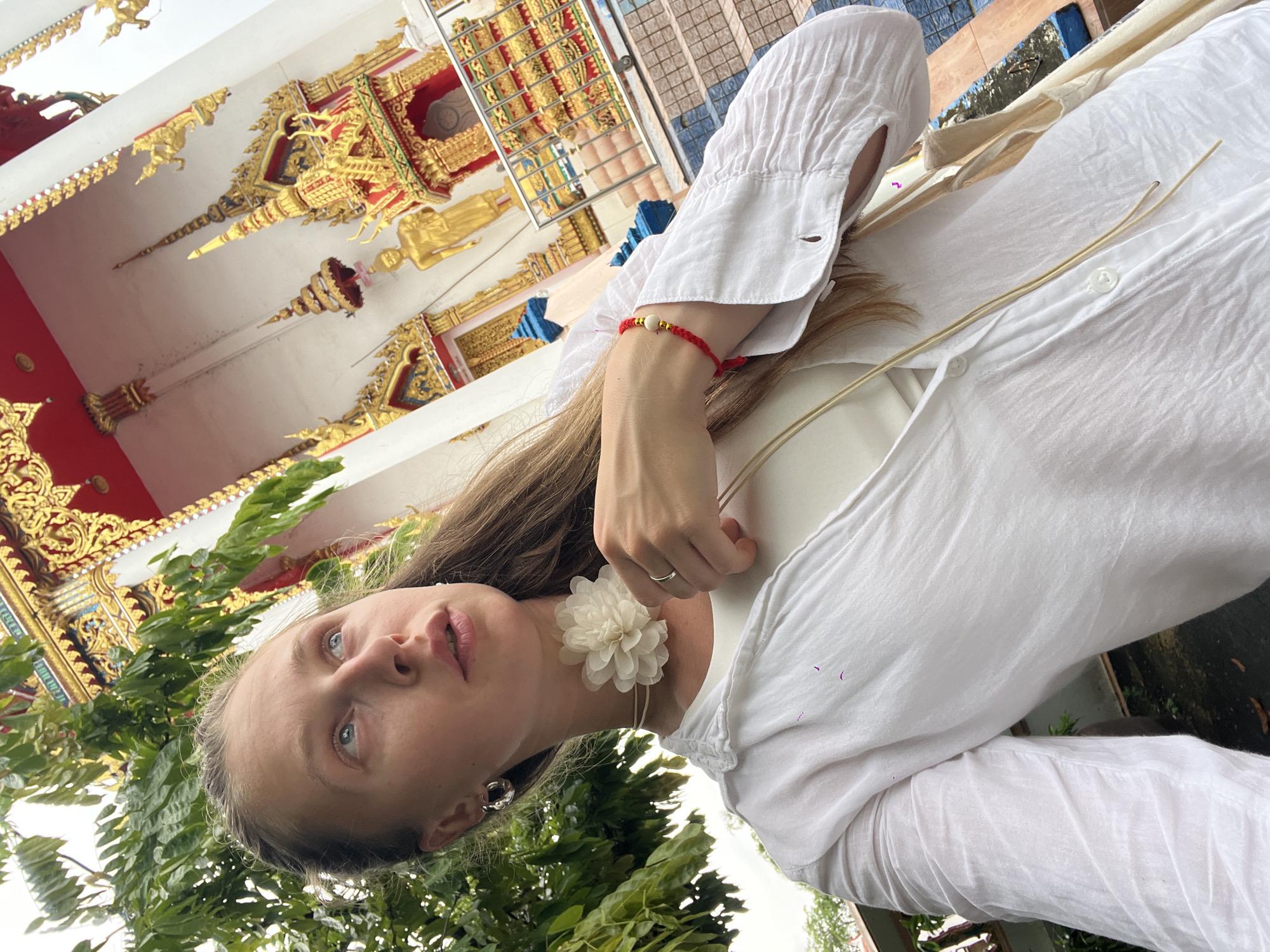 In Si Sunthon, Thailand, a young woman with light-colored hair pulled back stands in front of a temple. She is wearing a white button-down shirt over a white tank top, a white flower choker, a silver ring, and a red bracelet. The temple features ornate gold and red detailing, including statues and spires. Lush green foliage frames the scene from the left.