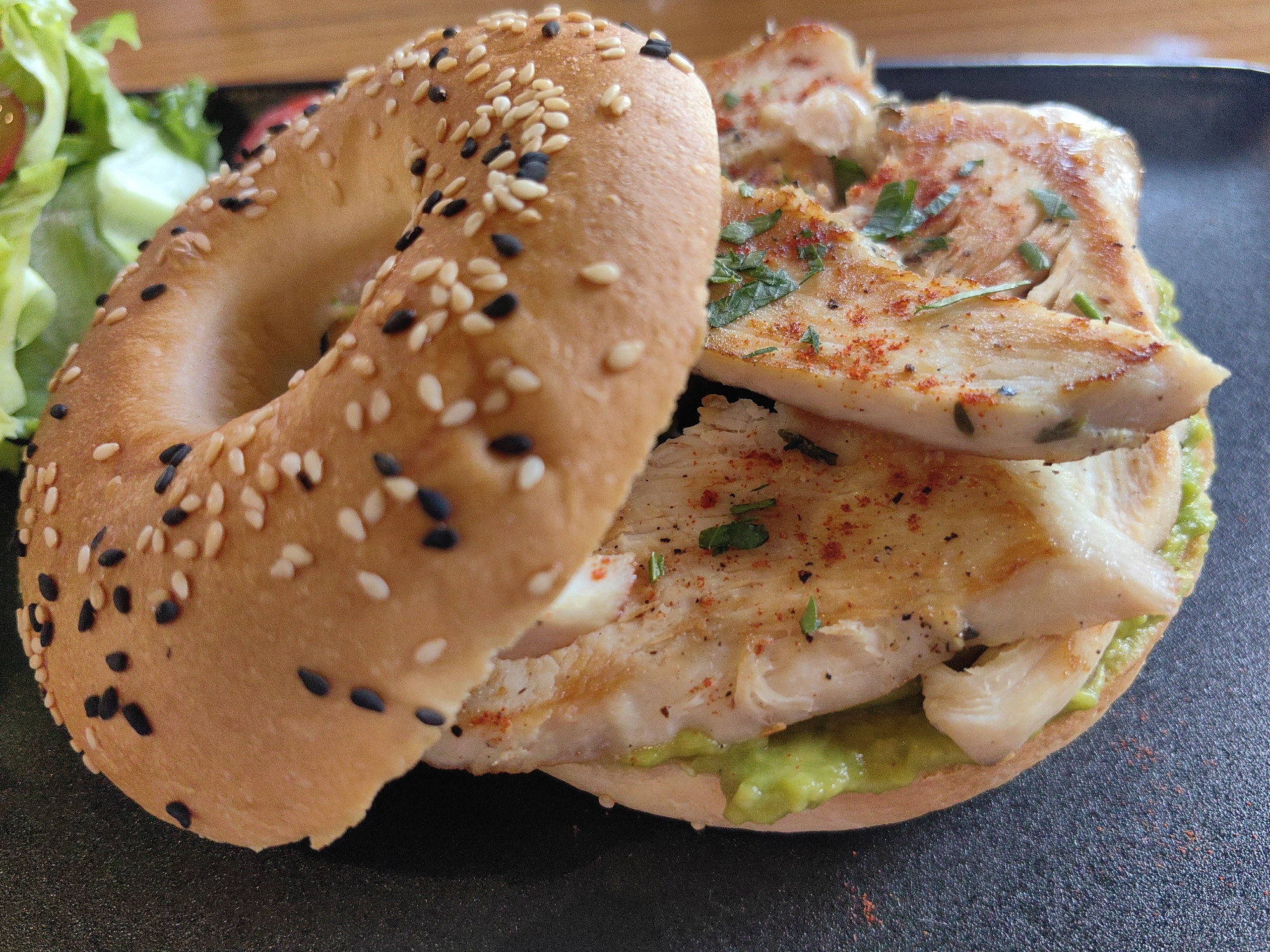 In Baan Bo Phut, Thailand, a close-up, eye-level shot captures a sesame seed bagel sandwich with grilled chicken, avocado, and a side of salad. The sandwich is presented on a dark plate, with the bagel, sprinkled generously with both white and black sesame seeds, forming the base. Slices of lightly seasoned grilled chicken, garnished with chopped herbs and red pepper flakes, are stacked within the bagel. A layer of vibrant green avocado mash is spread on the bottom half of the bagel. To the left, a portion of a fresh green salad with what appears to be cucumber and tomato is visible. The lighting is natural and bright, highlighting the textures and colors of the food. No people are present in the frame.