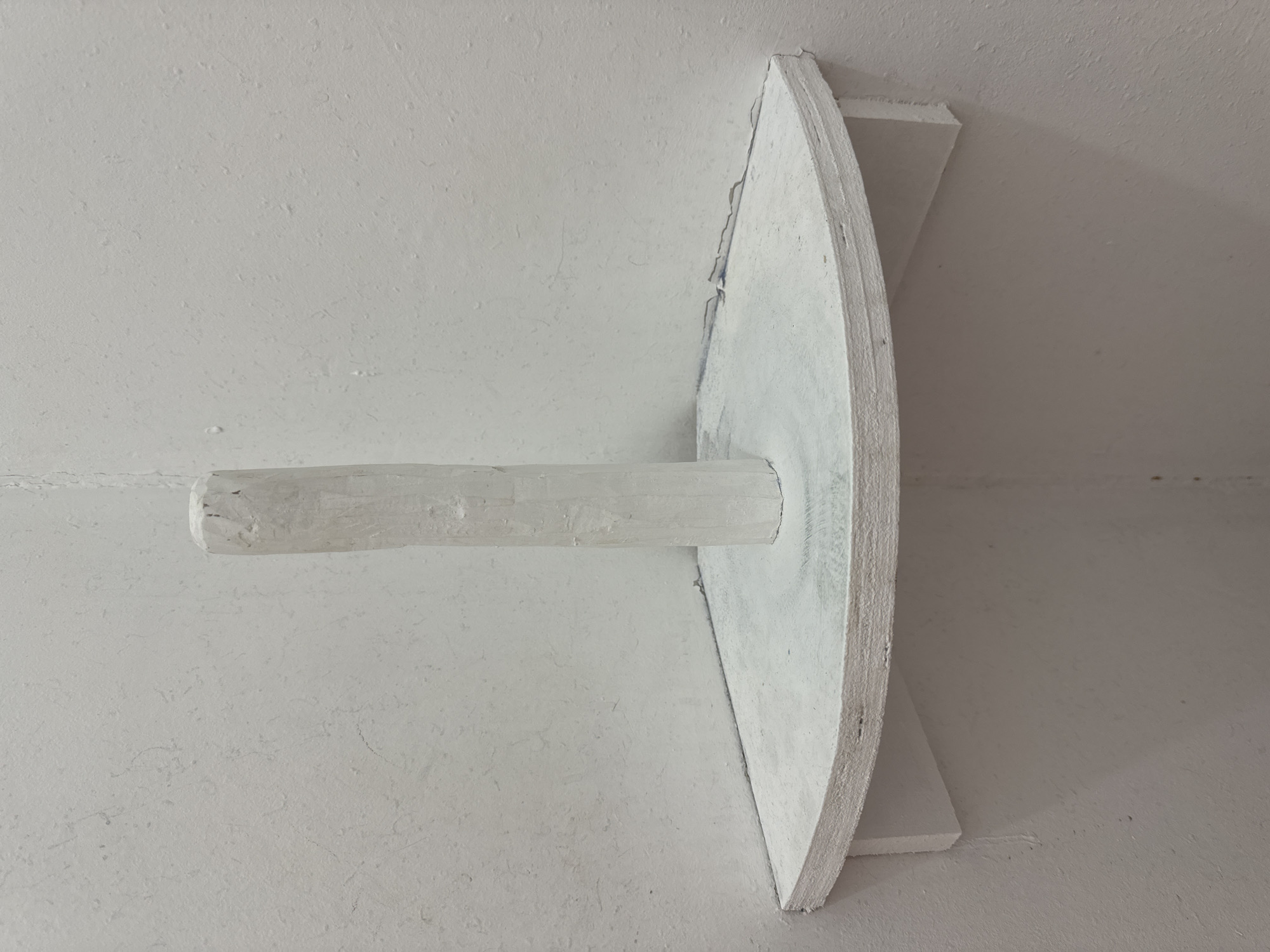 The image depicts a white corner shelf mounted on a textured white wall. A white, cylindrical object, resembling a piece of chalk or a small sculpture, stands vertically on the center of the shelf. The shelf itself is triangular with rounded edges and is supported by two white triangular brackets. The overall impression is one of minimalist art or a staged display within a room. There is no visible indication of its location within Waterford, Ireland.