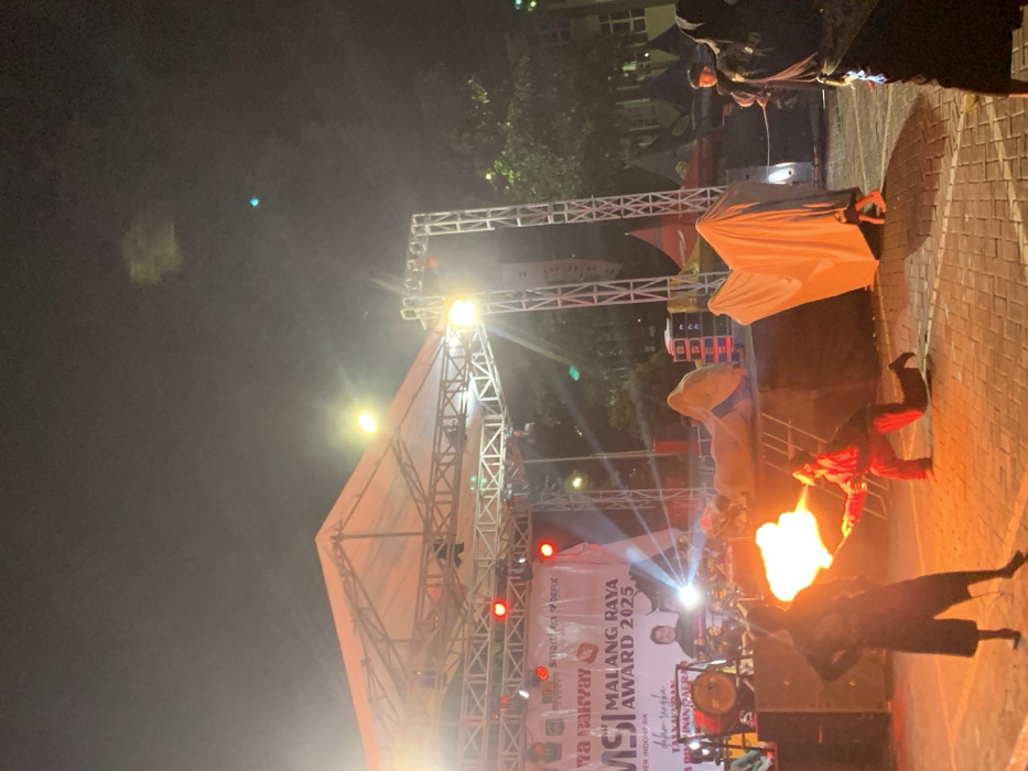 The scene takes place at night in Kecamatan Batu, Indonesia, at the coordinates -7.867, 112.514. There is a stage set up outdoors, with a large white canopy overhead and a metal truss structure holding up lighting fixtures. The stage has a large red sign that says "MALANG RAYA MSI AWARD 2025." There are several people on and around the stage, including a man playing a drum set, a man with a microphone, and a person in a black suit performing a fire-breathing act. Two people dressed in white sheets are standing to the right of the stage. The stage is set up in a paved area with a few trees and other buildings in the background. The scene is illuminated by stage lights and several other streetlights. The sky is dark with a few stars visible. The atmosphere is festive and celebratory.