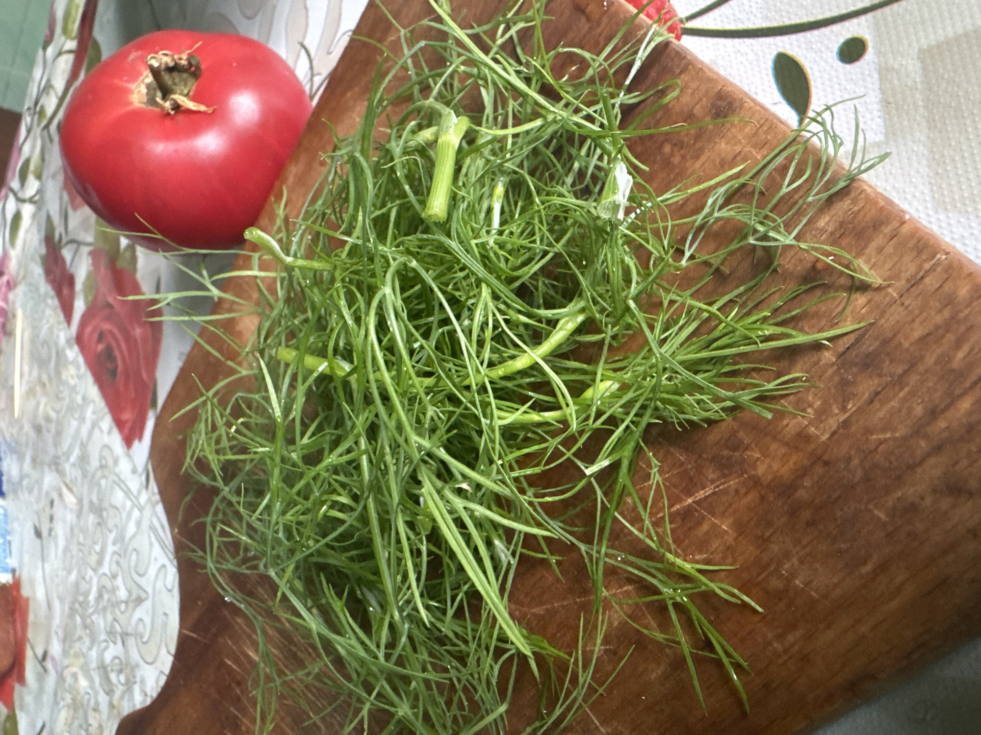 The image shows a wooden cutting board with a pile of fresh dill on it. To the right of the dill, there is a ripe red tomato. The cutting board is placed on a patterned tablecloth, which has floral designs in red and white. The scene appears to be in a kitchen or dining area. The image does not provide any information about the city of Красное, Russia, or any specific location within it. There are no people visible in the image.