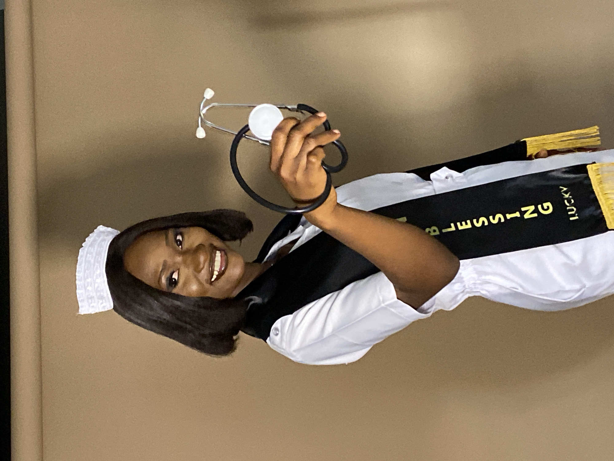 A young woman, possibly in her late teens or early twenties, is wearing a traditional white nurse's uniform with a cap. She is also wearing a black sash with gold fringe that reads "Blessing Lucky." She is holding a stethoscope in her hand. The background is a plain brown wall. The location is Gura Top, Nigeria.