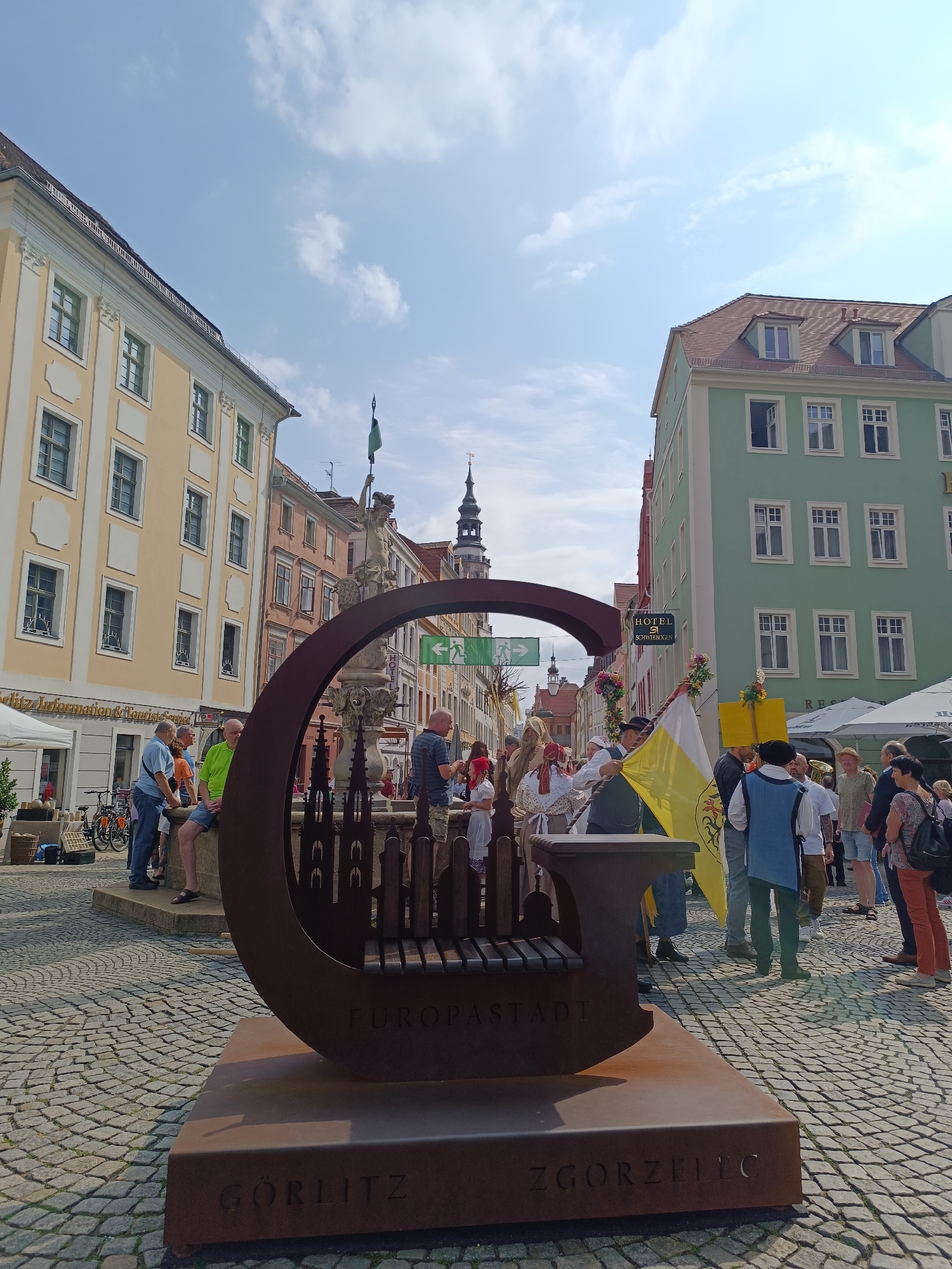 In the city of Görlitz, Germany, on a sunny day with a partly cloudy sky, a group of people are gathered in a cobblestone square. In the foreground, a large, stylized metal sculpture resembling the letter "G" serves as a bench. The sculpture is engraved with the words "EUROPATSTADT" on its seat and "GORLITZ ZGORZELEC" on its base. The sculpture is positioned in front of a Baroque-style fountain with a statue of a figure holding a green flag aloft.
Surrounding the fountain and sculpture are various buildings with colorful facades, characteristic of European architecture. One building to the left is light yellow with ornate white trim, featuring multiple windows. To the right, a building is painted a vibrant green, also with numerous windows. Other buildings in the background display shades of pink and orange.
A diverse group of individuals are present in the scene. Some are dressed in modern attire, while others are in historical costumes, suggesting a festival or historical reenactment. Several people are standing around, talking, and observing. A couple is seated on the sculpture, and others are near the fountain. In the background, a tent is set up, possibly for a market or information booth. The cobblestone pavement indicates an old town setting.
