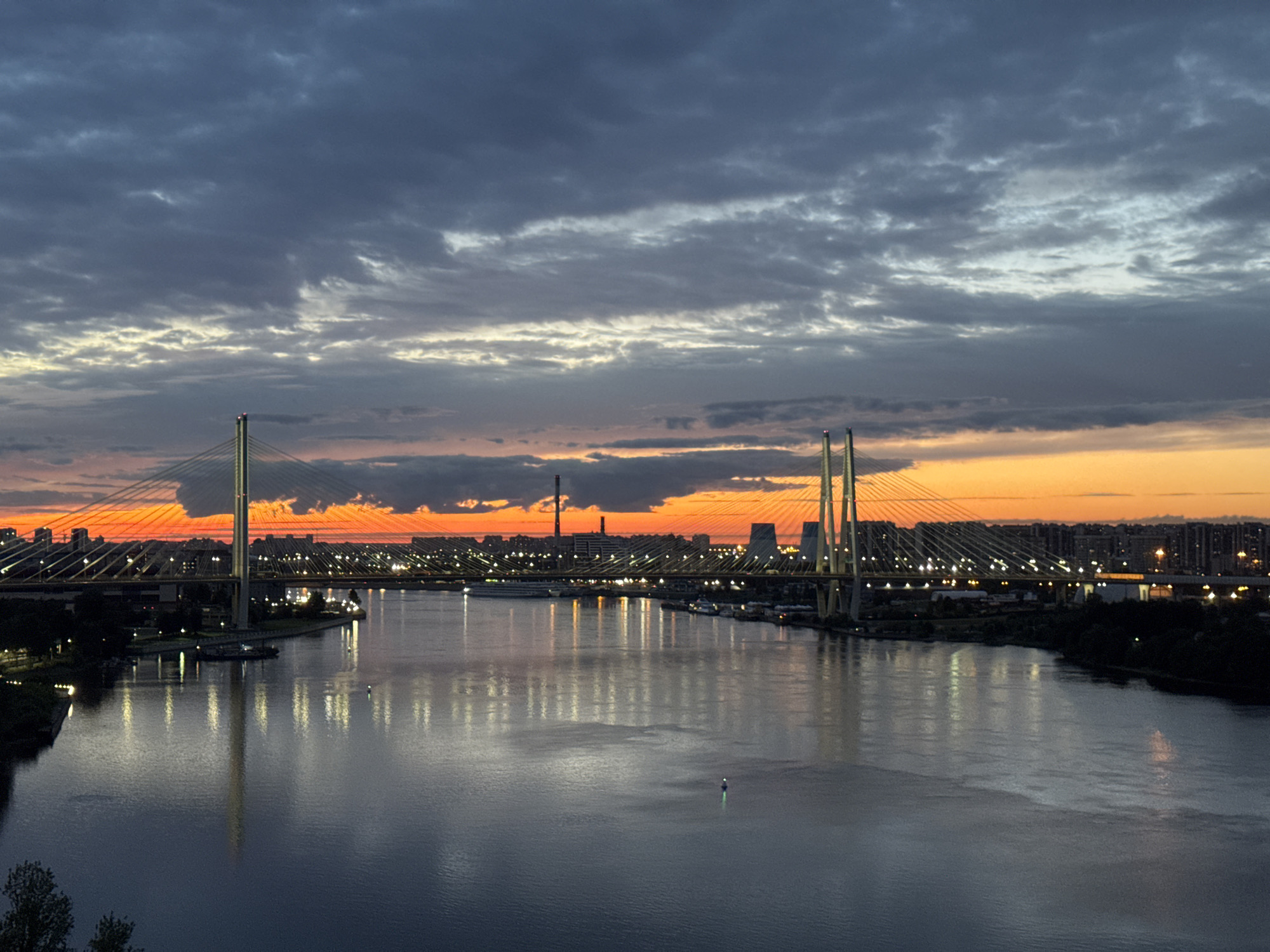 The image depicts a wide river flowing through an urban landscape during a sunset. The sky is filled with dramatic clouds, with vibrant orange and yellow hues near the horizon, transitioning to darker grays and blues overhead. Two prominent cable-stayed bridges span the river, illuminated by numerous lights. The bridges are supported by tall towers, and their cables are visible against the twilight sky. Along the riverbanks, there are buildings, streetlights, and dark silhouettes of trees. The water reflects the lights from the bridges and the city, creating a shimmering effect. A small, lit buoy is visible in the middle of the river. The overall scene suggests a twilight view of a major city with a prominent river and modern bridge infrastructure.