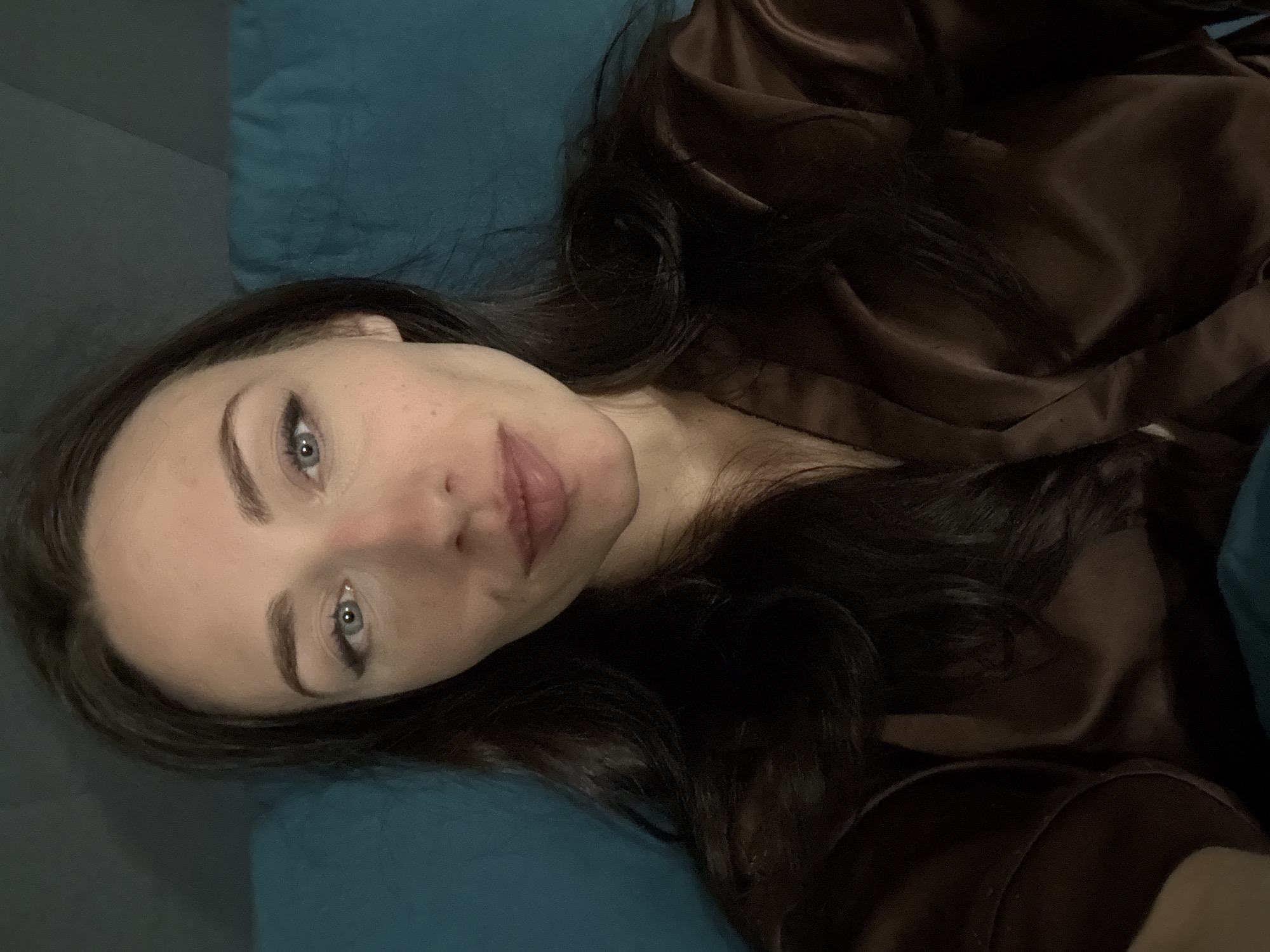 A woman with dark hair and light blue eyes is lying on a blue surface, possibly a bed or a couch. She is wearing a brown silky robe or top. The image appears to be a close-up selfie. There is insufficient information to determine her location in Saint Petersburg, Russia.