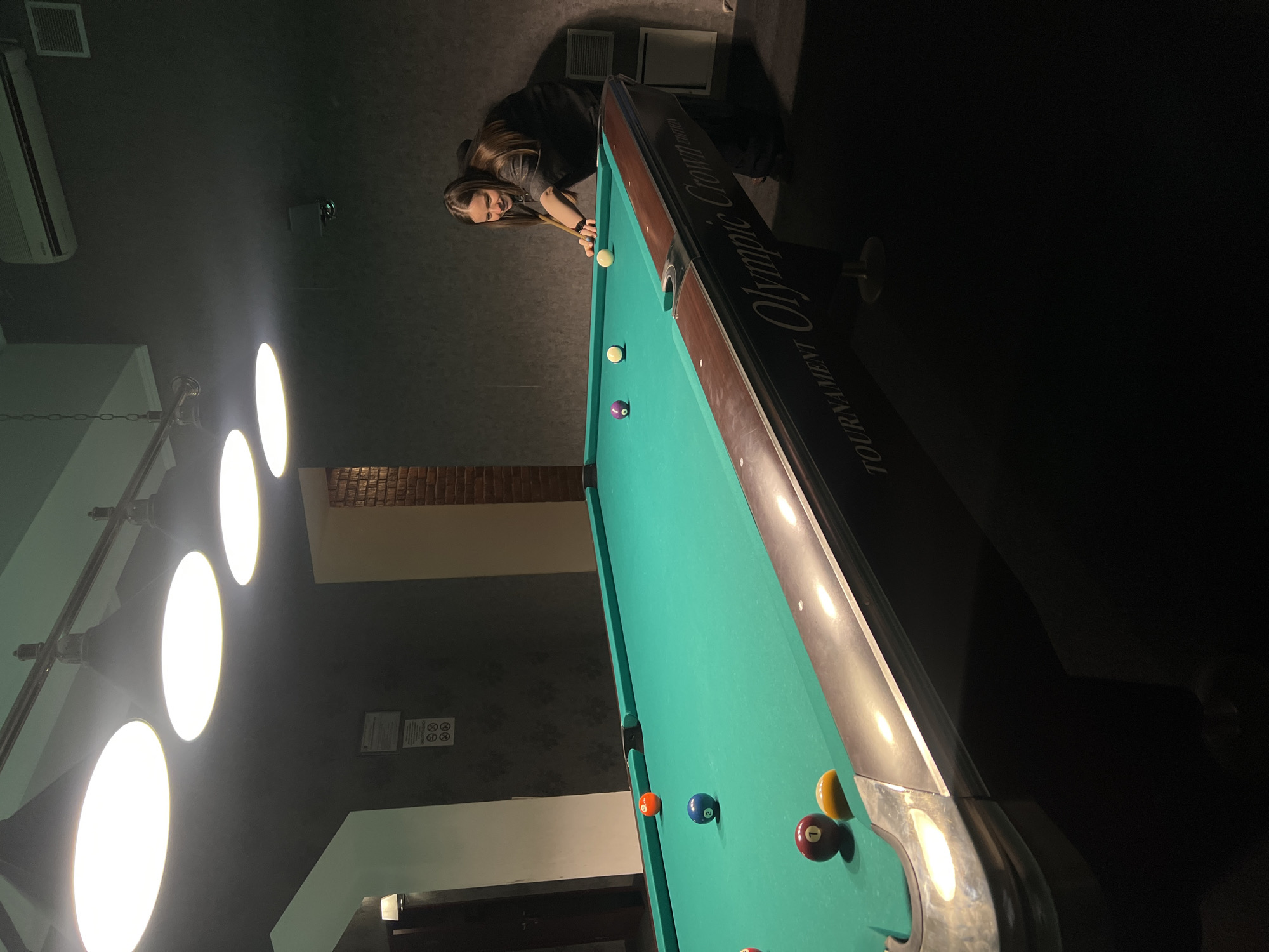 A low-angle, indoor shot depicts a billiards game in progress. A fair-skinned woman with long brown hair, wearing a grey t-shirt and dark trousers, is leaning over a green billiard table, cue stick in hand, preparing to strike the white cue ball. The table is of "Tournament Olympic Crown" make. Several numbered billiard balls are scattered on the table. Above the table, a series of round, bright lights are suspended. The walls are dark grey. A section of brickwork is visible behind the table. The location is presumed to be in Odesa, Ukraine, given the presence of the brand "Tournament Olympic Crown" which is associated with billiards tables from that region.