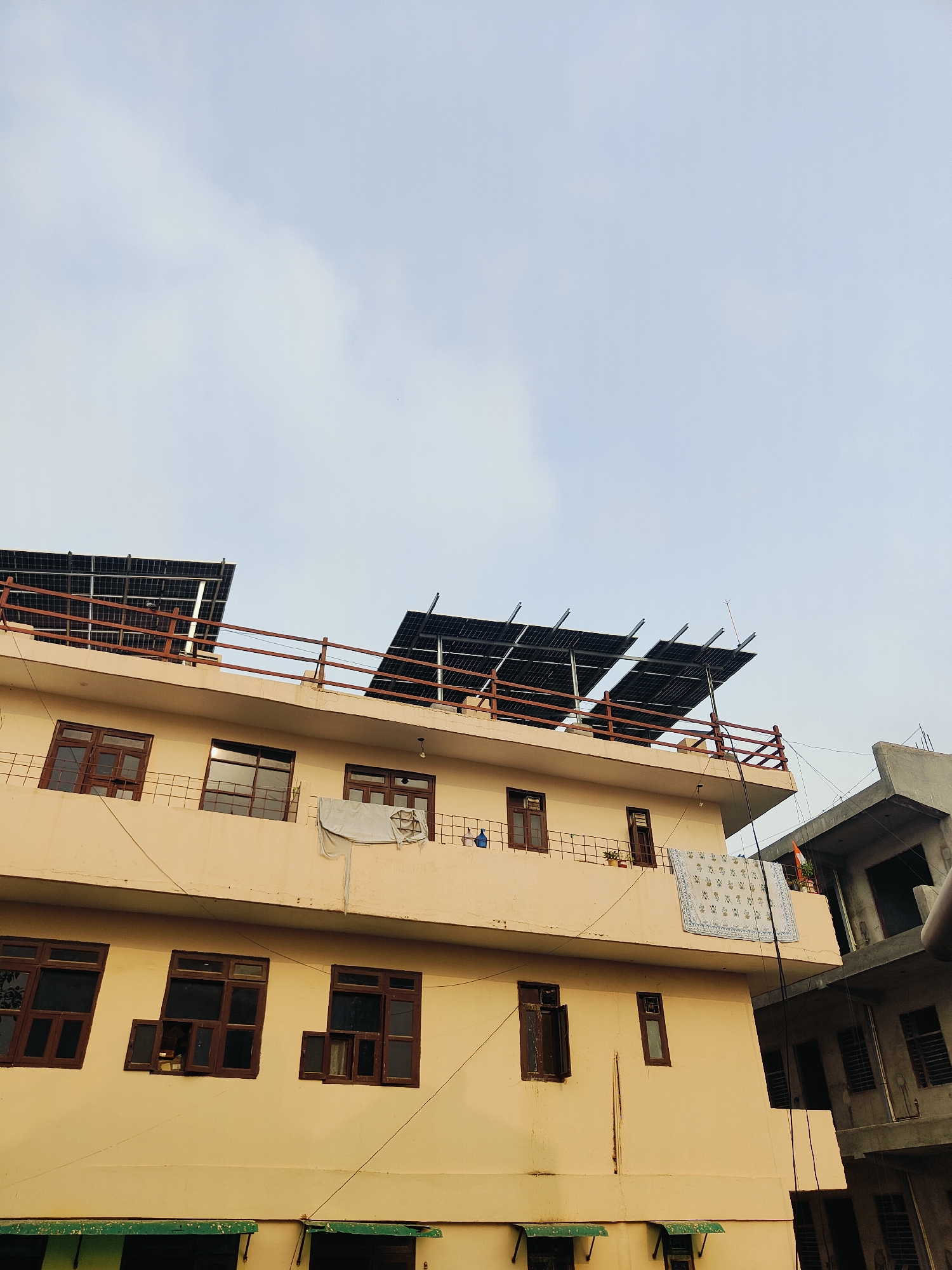 The image shows a multi-story building with a light yellow facade, possibly in Gurgaon, India. The building has several floors with multiple windows, many of which are open. On the rooftop, there are several solar panels installed, supported by a metal frame, and a reddish-brown railing. A white cloth with a patterned design is hanging from the railing on the second floor. Adjacent to the yellow building, a gray concrete structure is visible, appearing to be under construction or in a less finished state. The sky above is overcast with light clouds. There are no people visible in the image.