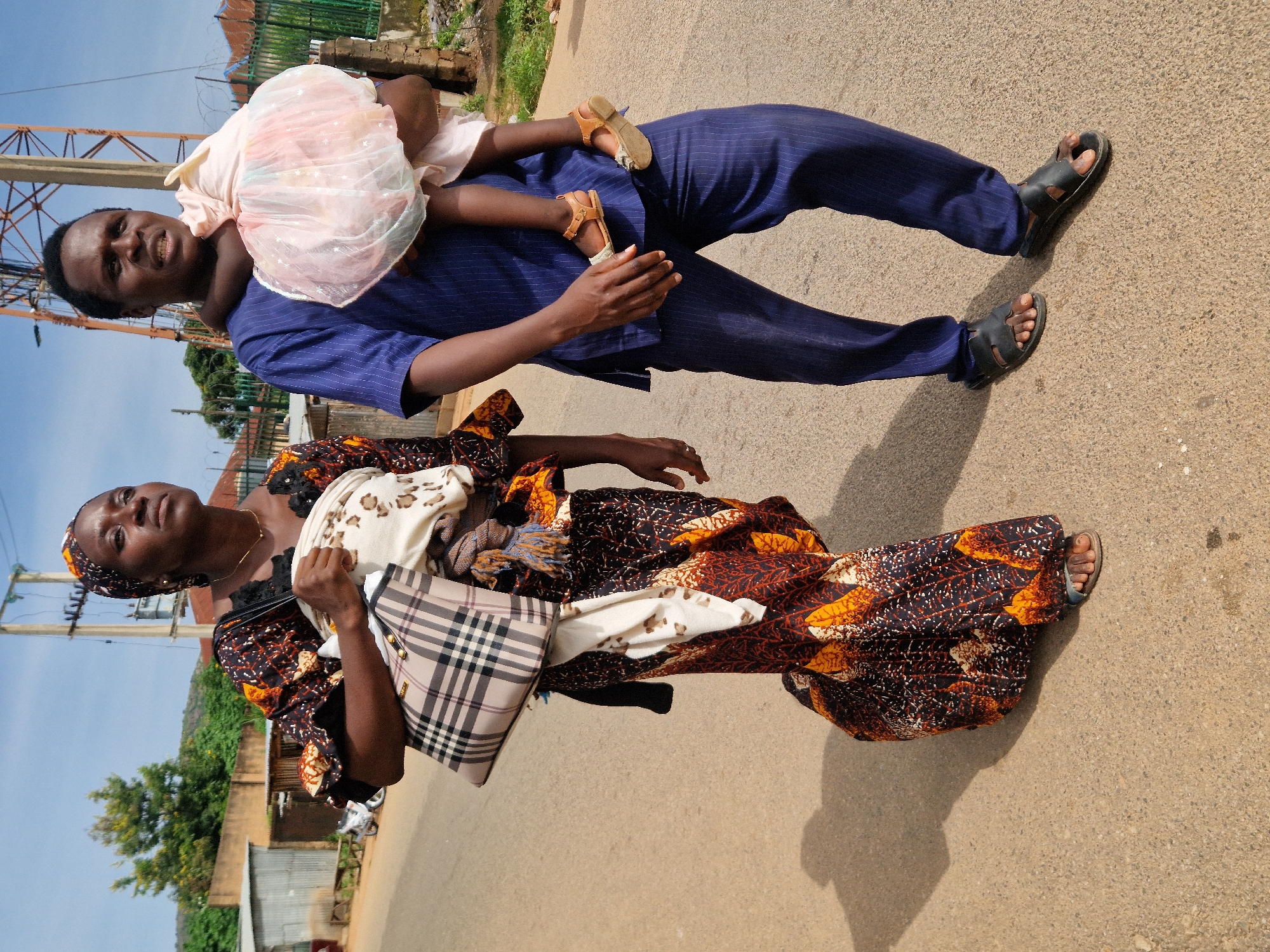 In Jos, Nigeria, a man and a woman walk on a paved road under a clear sky. The woman, dressed in a patterned brown and orange traditional dress and a headscarf, carries a beige and black plaid tote bag and a baby wrapped in a light-colored fabric over her shoulder. The man, wearing a blue pinstripe traditional outfit and black sandals, carries a young child in his arms, who is dressed in a light pink frilly dress and brown sandals. The background shows a few buildings, including a corrugated metal structure and a distant cell tower, with trees and a fence lining the right side of the road. The sun casts shadows on the road, indicating it is daytime.