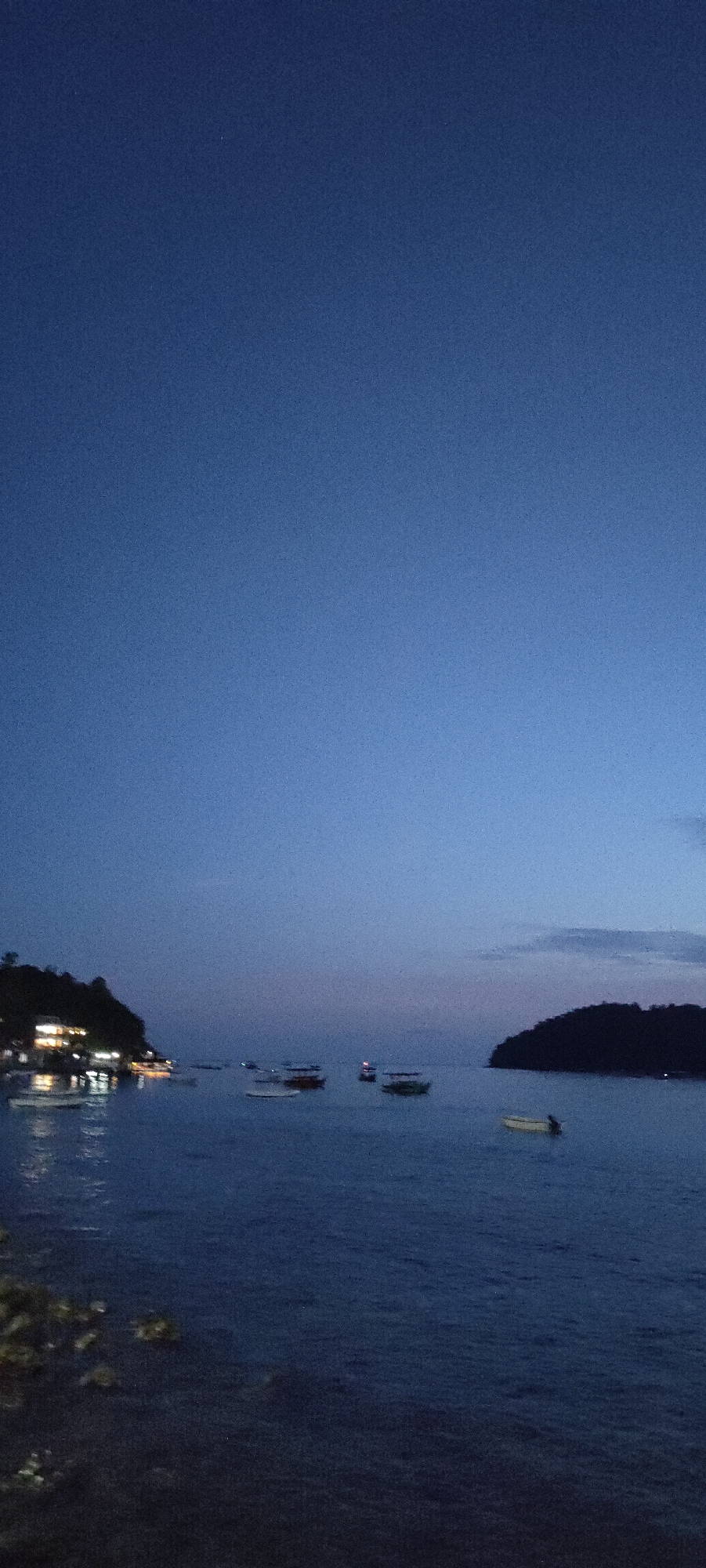 The image shows a body of water at dusk or night, likely in Sabang, Indonesia. Several small boats are visible on the water, some anchored and some appearing to be moving. In the background, a landmass with vegetation and lights from buildings is illuminated, suggesting a coastal settlement. The sky is a deep blue, transitioning to lighter shades near the horizon, and there are faint clouds. The overall atmosphere is calm and serene.