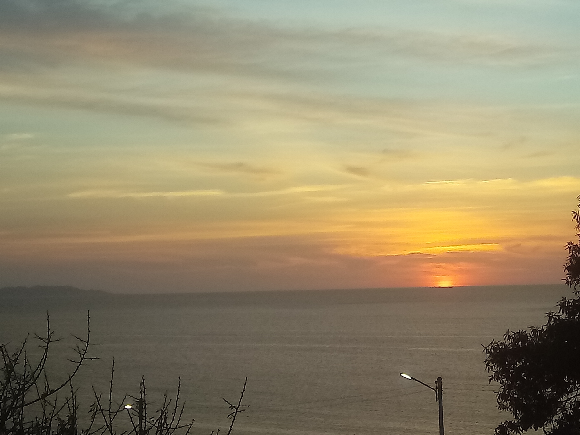 The image shows a sunset over the ocean in Juan Griego, Venezuela. The sky is a mix of blue, orange, and yellow, with the sun just above the horizon. A strip of land is visible in the distance. In the foreground, there are silhouettes of tree branches and a street light. The ocean is calm and reflects the colors of the sky.