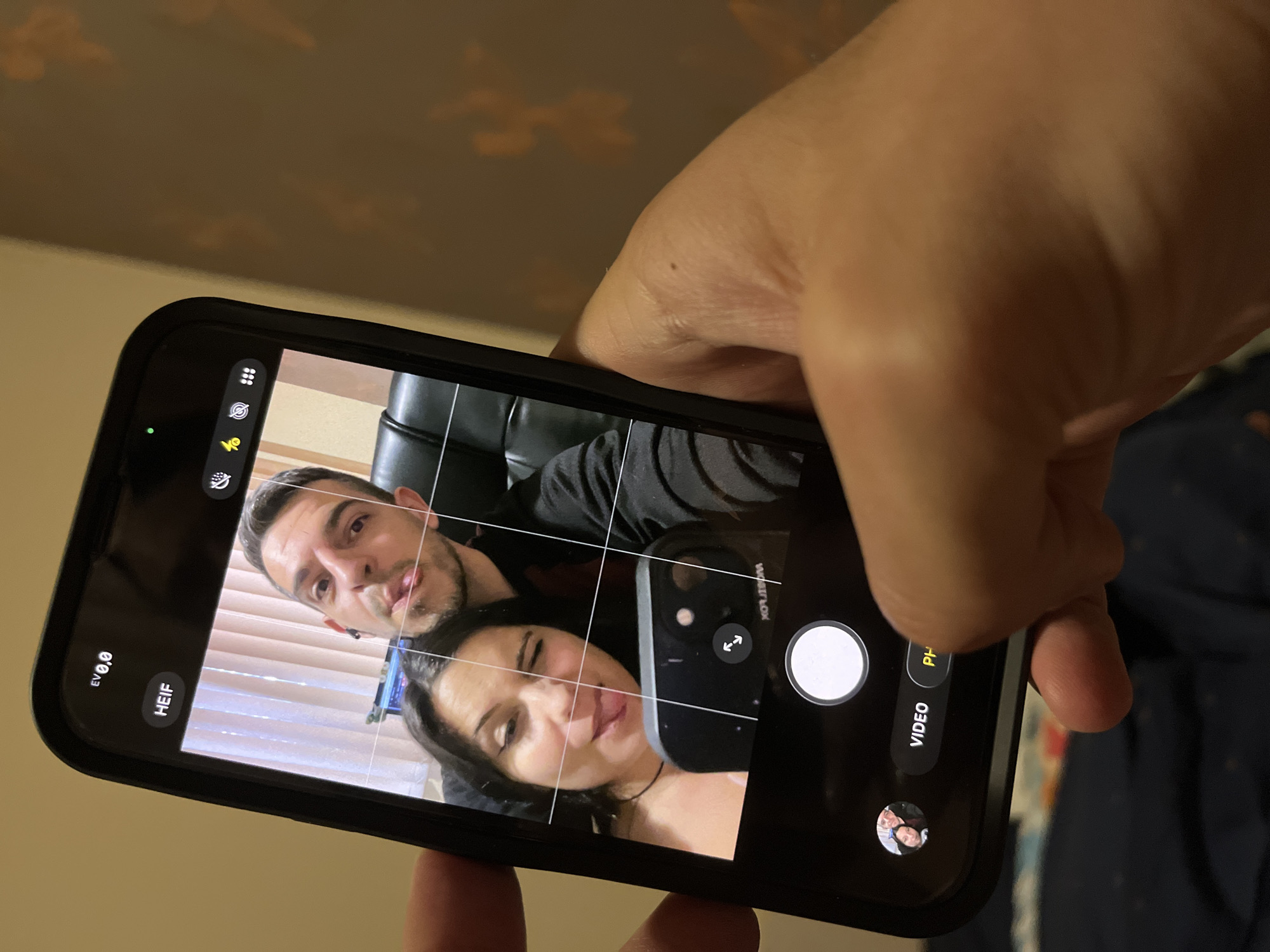 An iPhone, held by a person's hand, displays a selfie taken by a couple. The couple is positioned in the center of the screen, with the woman on the left and the man on the right. Both are making playful faces, with the woman winking and sticking out her tongue, and the man sticking out his tongue. The background shows vertical blinds and a dark chair. The phone's camera interface is visible at the bottom, showing options like "VIDEO" and "PHOTO". The location is the city of Sofia, Bulgaria.
