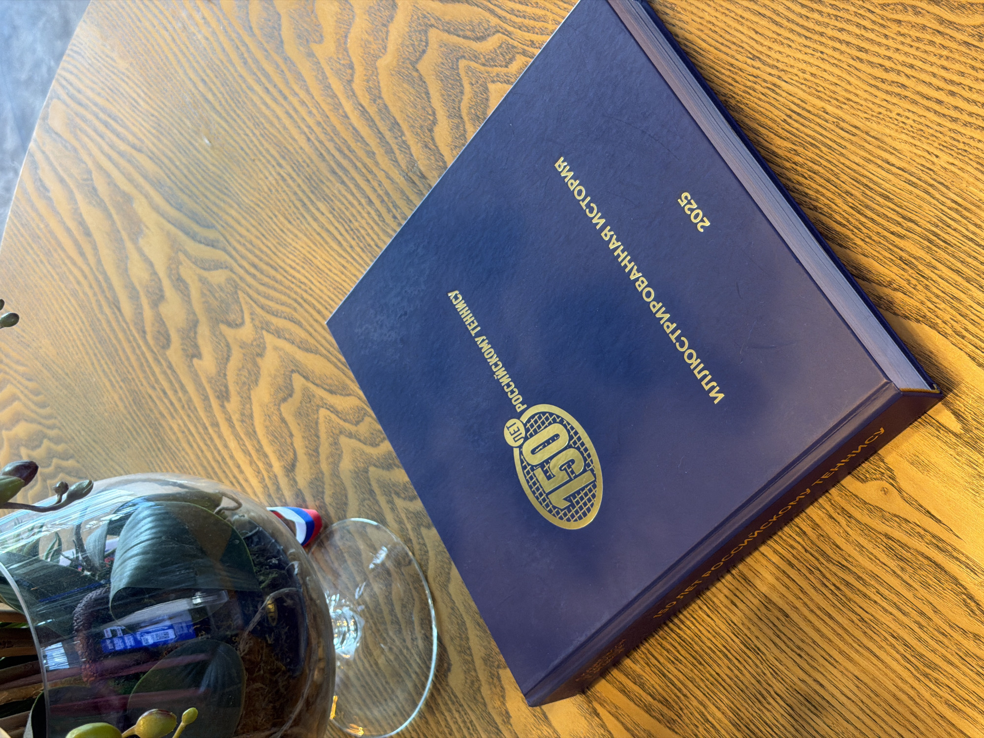 A close-up shot shows a hardcover book resting on a wooden table. The book is dark blue with gold lettering and a gold emblem, possibly a logo. Some text on the book is in Russian, with the year "2025" also displayed. Next to the book, a clear glass container, possibly a terrarium or decorative vessel, holds plants and other items. Part of a glass from what appears to be a drinking glass is visible next to the container. The wooden table has a light brown hue and a noticeable wood grain pattern. The setting appears to be indoors. It is not possible to confirm this is taken in Ovoshchnoy, Russia, based on the image alone.