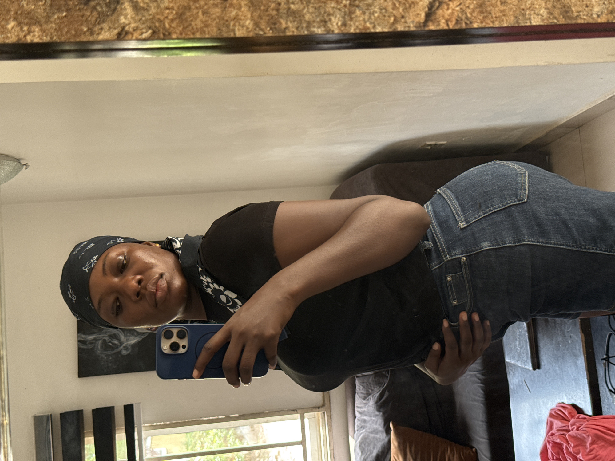 A dark-skinned person wearing jeans and a dark headscarf is taking a mirror selfie with a blue smartphone. The background reveals a room with a window and a sofa. The location is likely indoors, based on the domestic setting. Specific location in Abuja, Nigeria cannot be determined from the image.