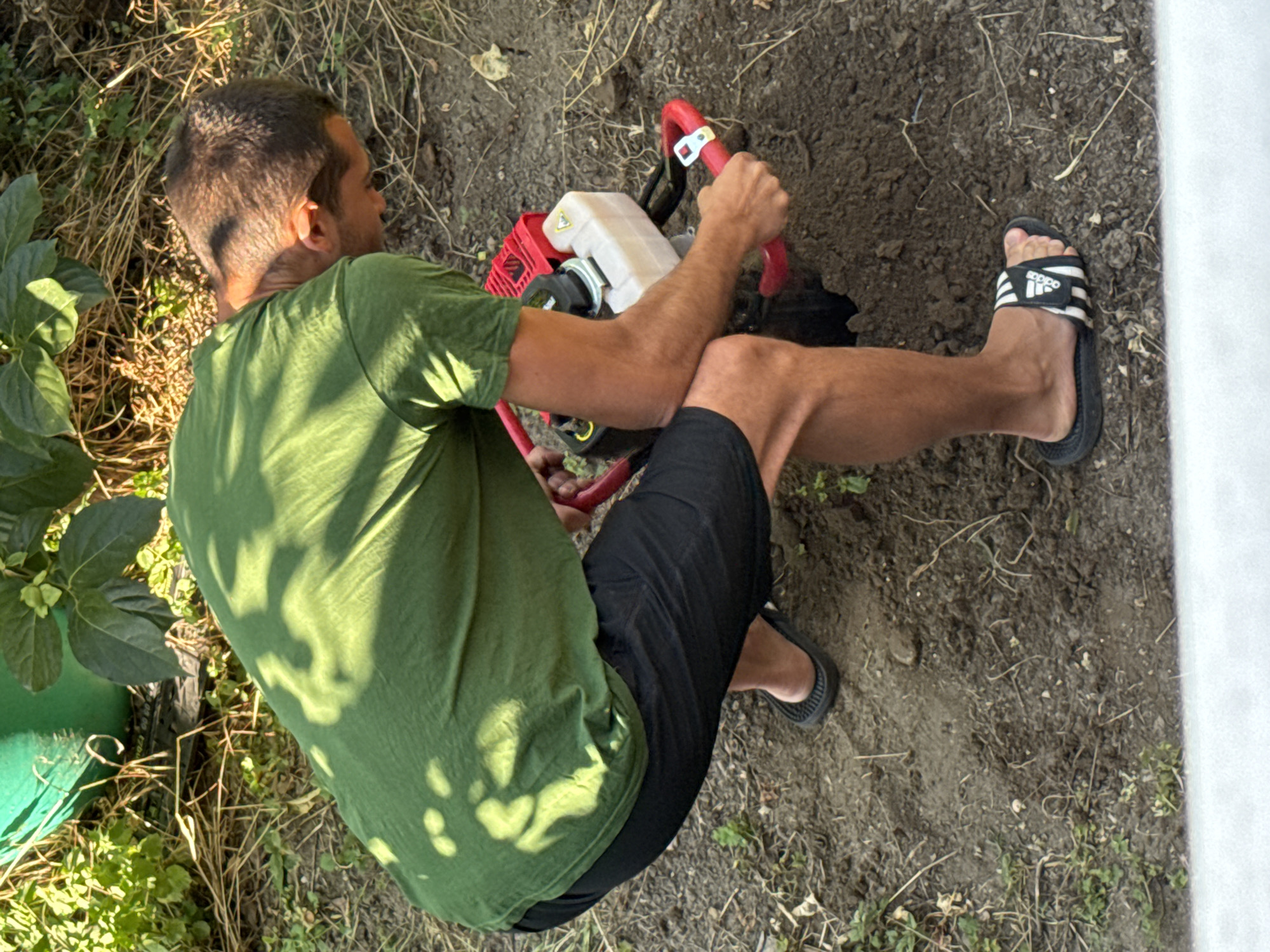 A male, possibly in his late 20s to early 40s, with short brown hair, is crouched down in a garden area, operating a red and black gas-powered soil auger. He is wearing a green t-shirt and black shorts, with black Adidas slide sandals on his feet. The ground is covered in loose soil, dry grass, and some small green plants. In the background, a large green plastic pot is partially visible, along with leafy green plants. The scene suggests an outdoor activity, likely gardening or landscaping, taking place in a residential or community garden setting within Plovdiv, Bulgaria. Sunlight casts dappled shadows on the man's t-shirt and the surrounding ground.