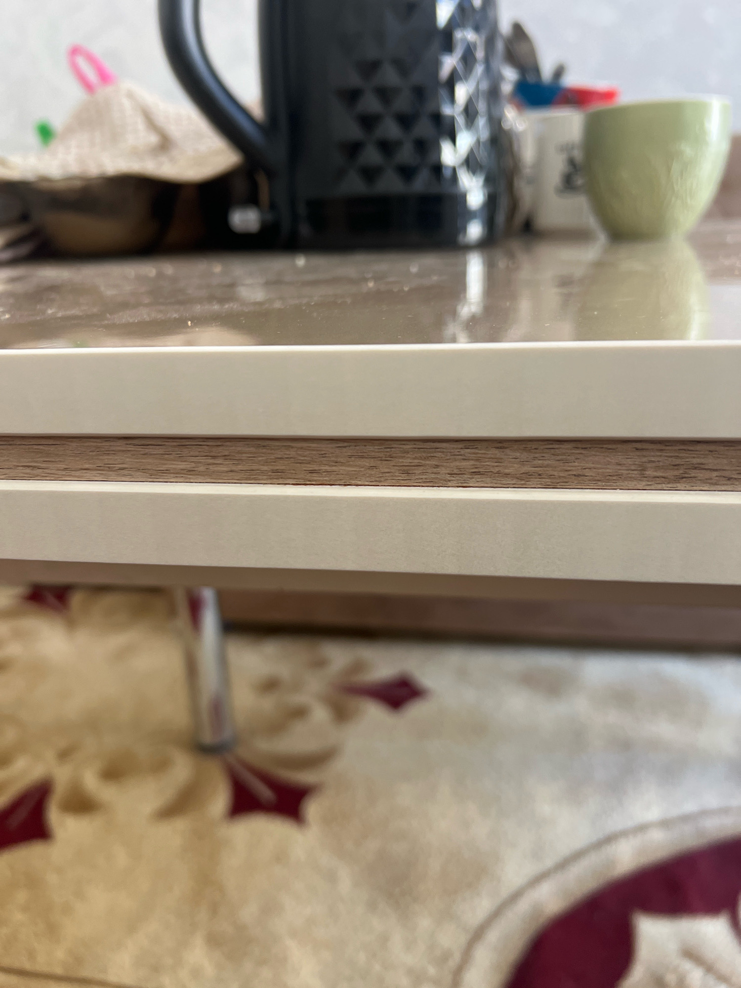 The image shows a close-up of a table with a light-colored surface and a wooden inlay. In the background, a black electric kettle is visible, along with a light green mug and other kitchen items. The table is positioned over a carpet with a beige and red floral pattern. A metallic leg of what appears to be a chair or table is also partially visible in the lower-left corner. The scene is indoors, likely in a kitchen or dining area, in an unknown city and location.