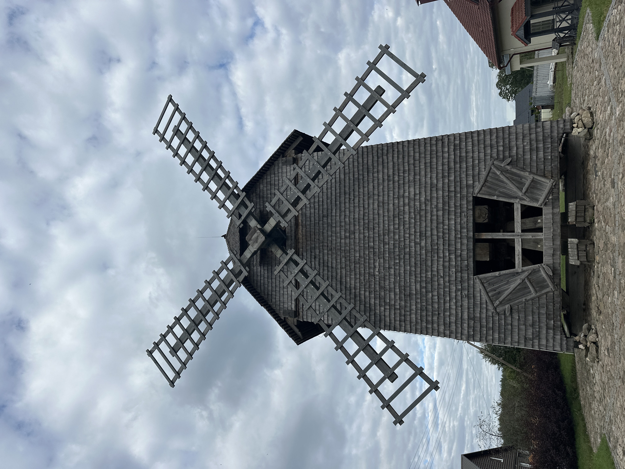 A wide-angle, low-angle, full frontal view shows a weathered, wooden Dutch windmill against a cloudy sky. The windmill is constructed from dark, overlapping wooden shingles, covering its conical structure. Four large, wooden, lattice-style windmill blades extend outward from the top of the structure. The blades are unfinished wood and appear to be weathered. Two large, wooden doors are open at the base of the windmill, revealing a dark interior. The windmill rests on a foundation of gray, irregular stones. The ground in front of the windmill is paved with cobblestones. To the left and right of the windmill are glimpses of other buildings and foliage. On the far left, a dark wooden house with a white-trimmed window is partially visible. Behind the windmill, there is a dense, dark red bush and a line of trees. To the right of the windmill, a building with a red tile roof and light-colored walls is visible, along with a small, dark wooden structure with a peaked roof. The sky is overcast with numerous gray and white clouds. The location is identified as Naroch, Minsk Region, Belarus.