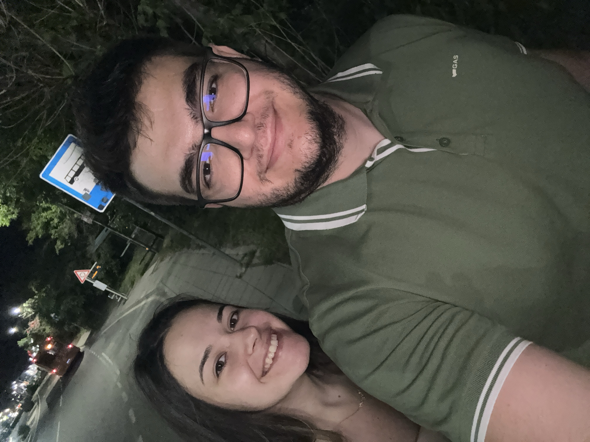 Two young adults, a woman and a man, are captured in a selfie taken outdoors at night on an urban street in Sofia, Bulgaria. The woman, positioned on the left, smiles broadly, displaying her teeth. She has long dark hair and wears a light-colored top with a delicate gold-colored necklace. The man, positioned centrally and to the right, also smiles, wearing black-framed glasses that reflect ambient light. He has a beard and mustache and is dressed in a dark olive green polo shirt featuring white and green striped cuffs and collar, with a small "GAS" logo on the left chest.
The background reveals a street scene at night. A paved road with white dashed lane markings is visible to the upper left, where a bus with illuminated red taillights is seen moving away in the distance. A sidewalk or pedestrian path runs alongside the road. Prominent street furniture includes a blue square road sign with a white bus icon, denoting a bus stop, located above the man's head. Further in the background, a red triangular warning sign with an exclamation mark is visible, alongside what appears to be a bus stop shelter and other street poles. The area is lined with trees and bushes, particularly on the right side, and is illuminated by multiple streetlights, creating a distinct night environment with visible light sources and reflections. The subjects appear to be interacting casually, likely posing for the photograph together.