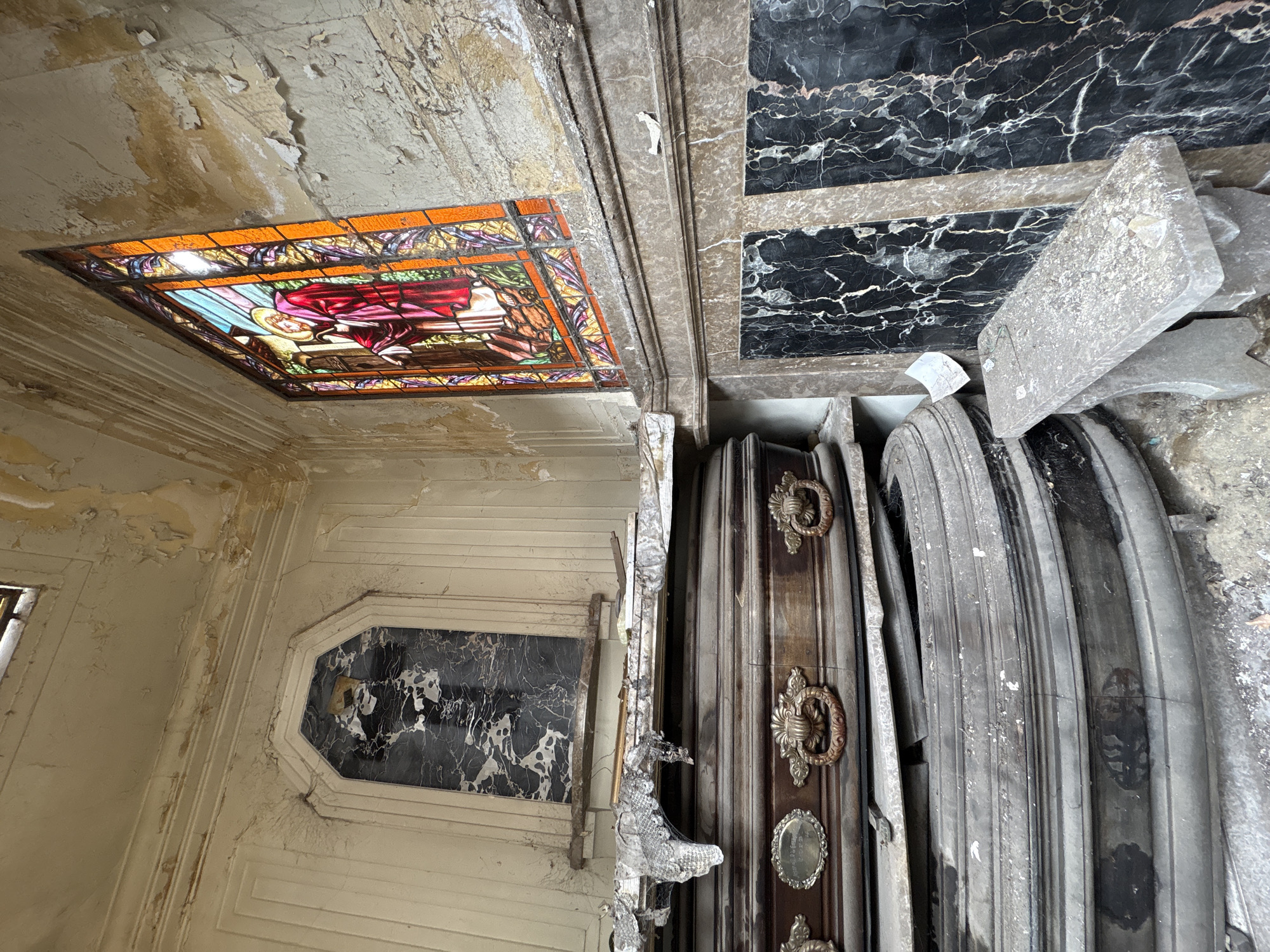 The image captures the interior of a neglected mausoleum or crypt within La Recoleta Cemetery in Buenos Aires, Argentina. The room's light-colored walls and ceiling exhibit extensive paint peeling and discoloration. A prominent vertical stained-glass window on the right depicts a haloed figure in a red robe, consistent with Jesus, knocking on a door; the glass features rich hues of red, blue, orange, and green. The lower walls are paneled with black marble displaying white veining, capped by lighter marble trim.
To the left, multiple wooden caskets are stacked vertically. The visible upper caskets are dark, ornately carved, and equipped with brass ring handles and an oval brass plaque; they are heavily obscured by dust, cobwebs, and draped with a deteriorated white lace-like fabric. Below these, additional dark, rounded caskets or urns are discernible, equally covered in grime and appearing structurally compromised. Above the stacked caskets, a recessed rectangular niche on the back wall holds a dark marble slab. A small, rectangular gray stone bench, also covered in dust and scattered debris, occupies the lower right foreground. The entire environment is characterized by significant dust accumulation and general disrepair, indicating a prolonged state of neglect.