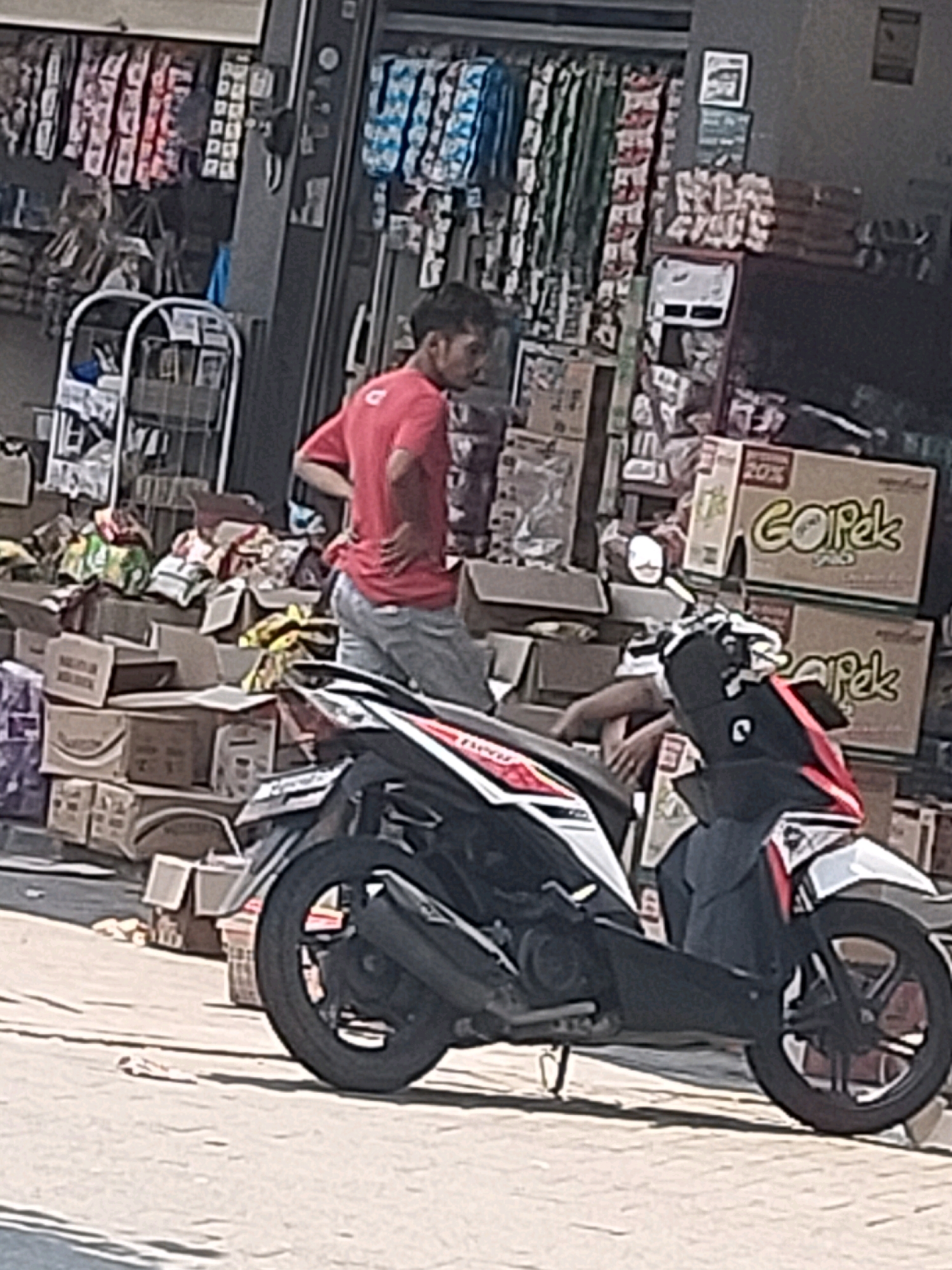 In Panjerejo, Indonesia, a man in a red shirt and blue jeans stands by a scooter, his right hand on his hip. He is facing left, his head turned slightly to the right, observing something out of frame. The scooter, predominantly black with red and white accents, is parked on a paved surface. Behind the man, a store displays various goods, including boxes stacked high and items hanging on racks. The scene suggests a commercial area, possibly a street-side shop.