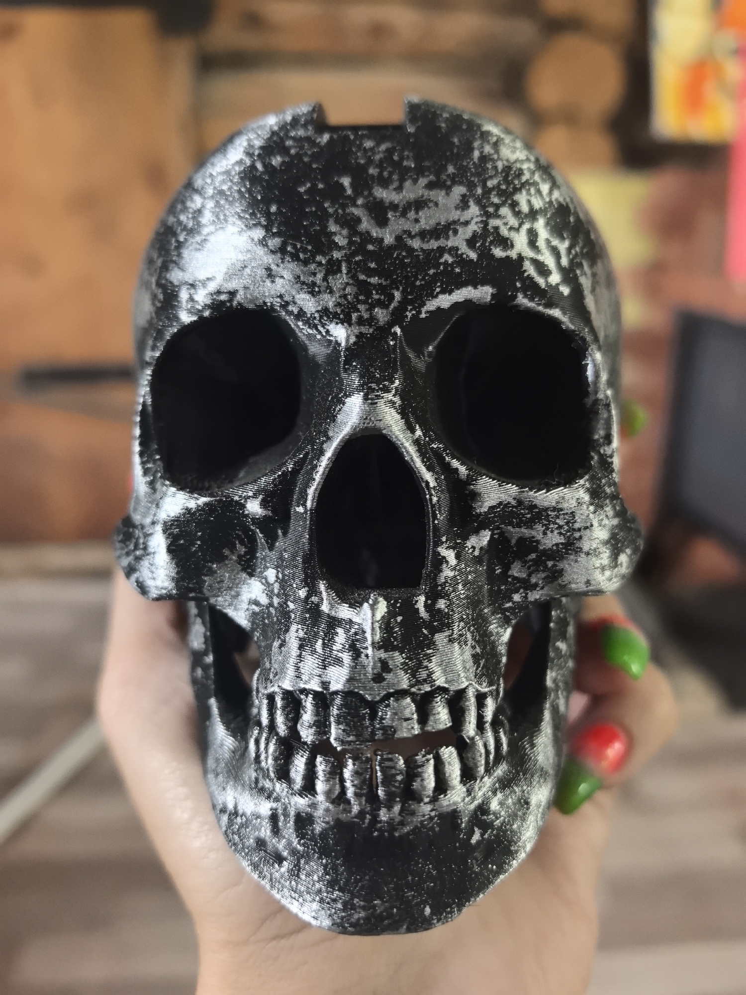 A hand holds a 3D-printed skull painted in black and silver with a rough texture. The skull's eye sockets and nasal cavity are dark, and the teeth are visible. The fingernails on the hand are painted with a green and red gradient. The background is blurred, showing a brick wall and a glimpse of a computer screen. The location in the city of Noviy, Russia is unknown as it is not apparent from the image.