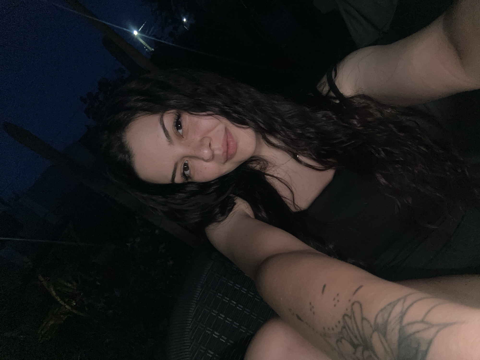 A woman with dark, curly hair smiles at the camera. She is wearing a black top and has a tattoo on her arm. The background is dark, with some trees and a streetlight visible. The location is Skala, Bulgaria.