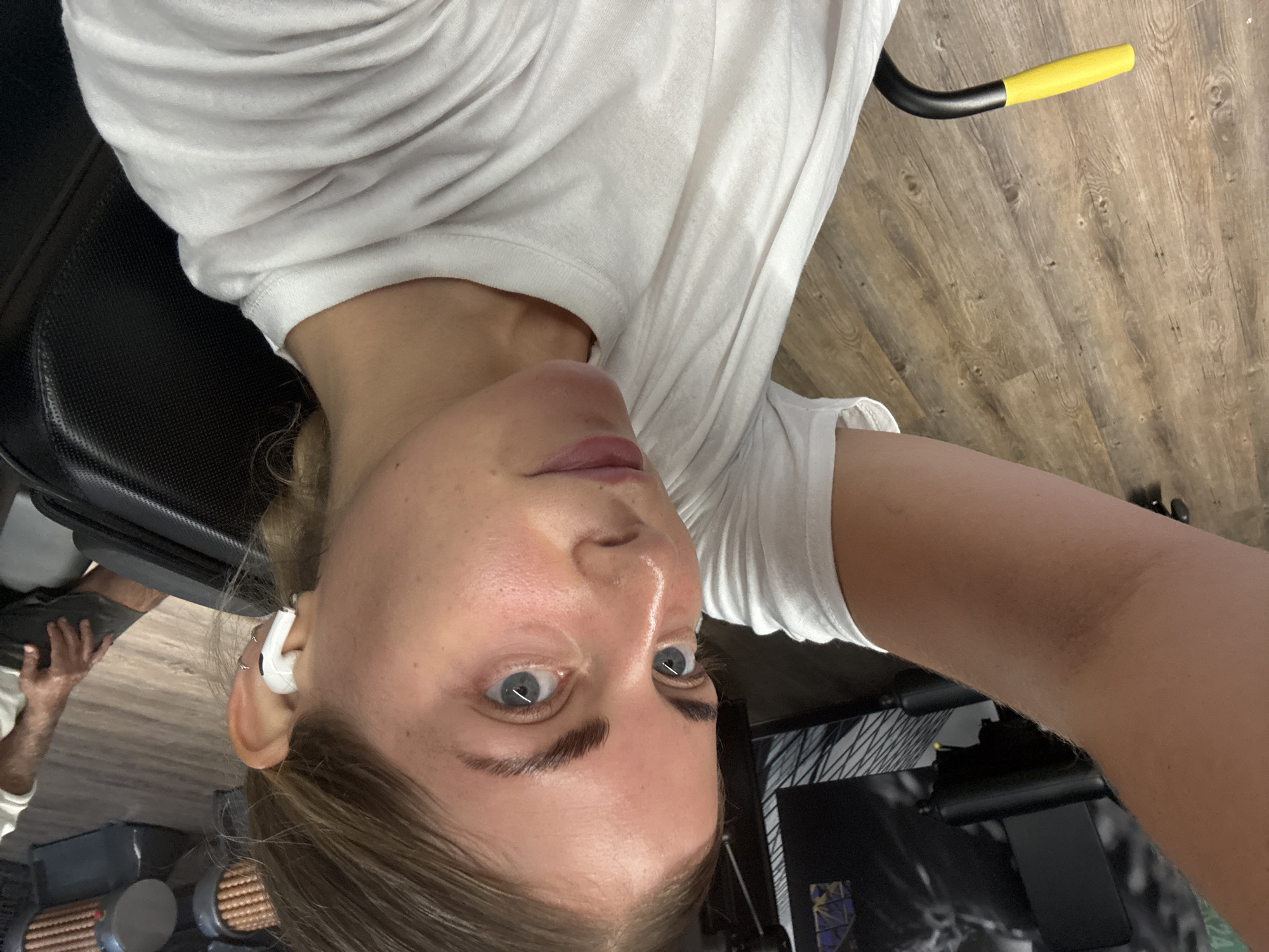 A selfie taken in a gym with wooden floors. A woman with fair skin, brown hair, and blue eyes is wearing a white T-shirt and a white earbud in her left ear. In the background, there is gym equipment including a black bench and yellow handle. Part of a man's leg is visible in the lower left corner. The location in the city of Paris, France cannot be determined.