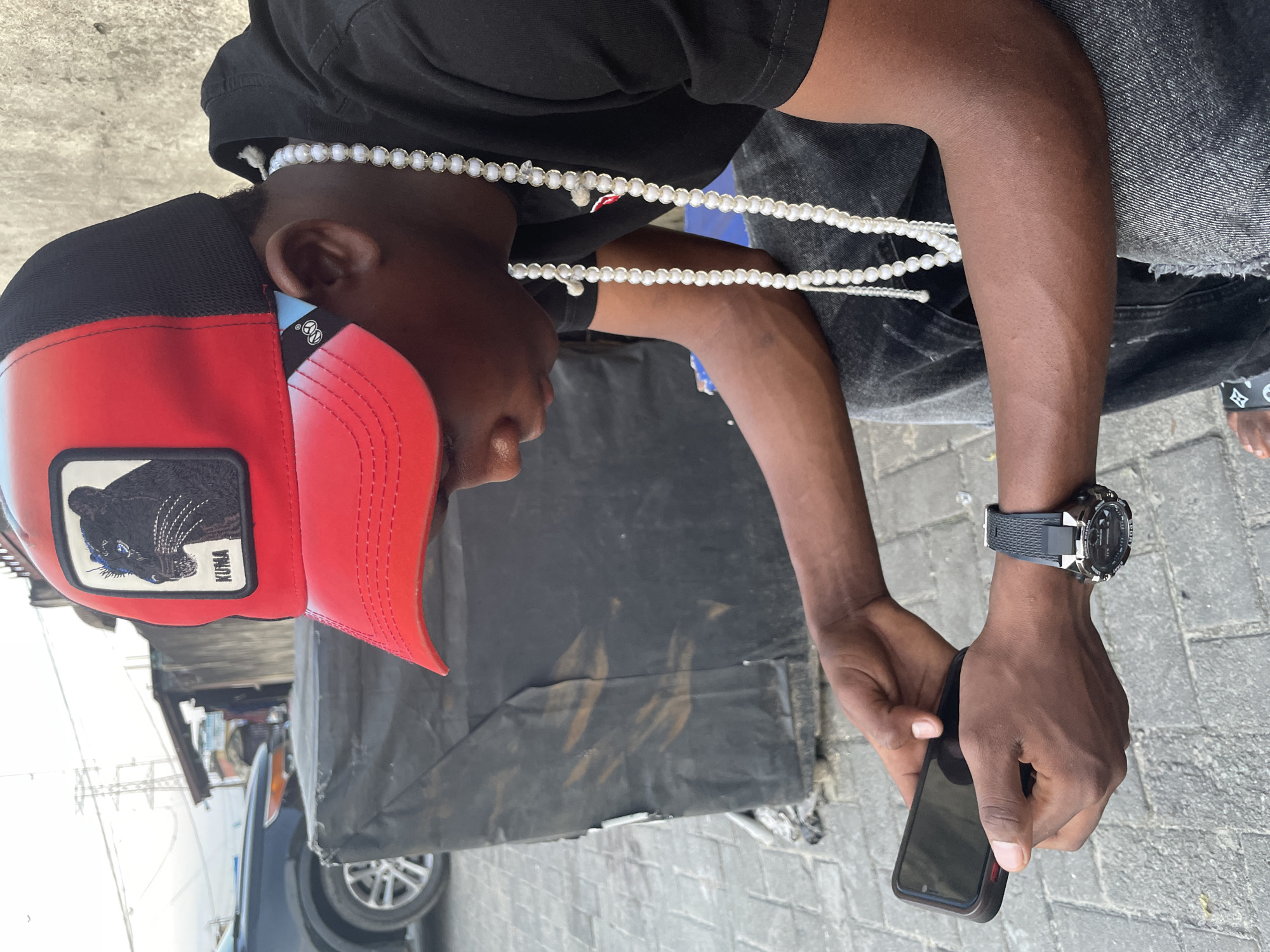 A young Black man, wearing a black t-shirt, black jeans, a red and black cap with a black panther logo, and a pearl necklace, sits outside. He is holding a black smartphone in his left hand and looking down at it. A black digital watch is on his right wrist. The man appears to be in Ado, Nigeria, indicated by the surrounding environment which features a paved ground with interlocking bricks, a large black tarp-covered object, a dark-colored car, and a distant power line tower. The man's face is partially obscured by his cap and the angle of his head, but his hands and arms are clearly visible, along with his watch and necklace. The lighting suggests it is daytime, possibly late afternoon or early evening, with overcast skies.