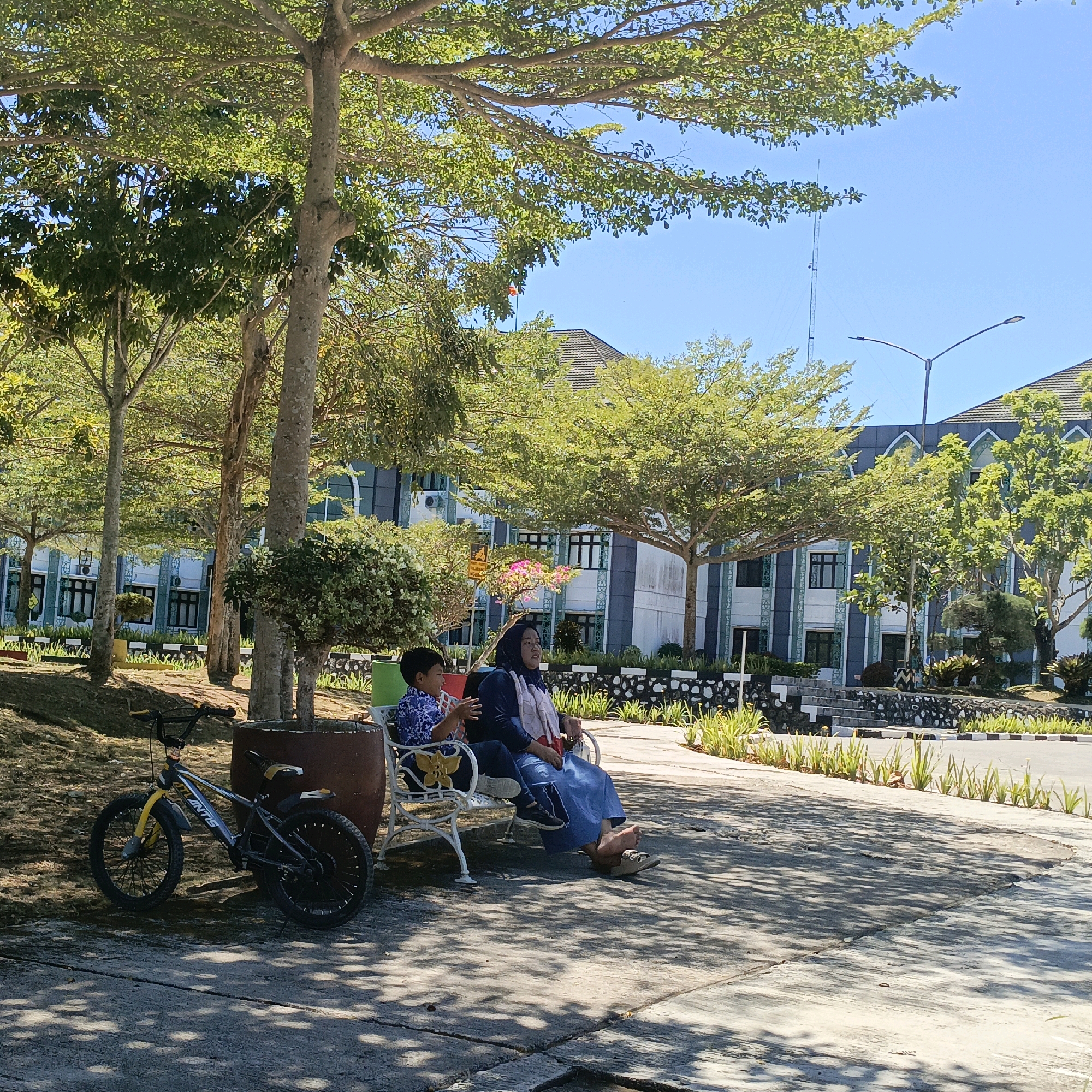 The image depicts an outdoor scene in Salatiga, Indonesia. In the foreground, a bicycle is parked on a concrete surface. A white bench is occupied by two people, a woman wearing a headscarf and a blue dress, and a young boy in a blue patterned shirt. They are sitting under the shade of a large tree with lush green leaves. In the background, a modern building with a patterned facade and arched windows is visible. The sky is clear and blue, indicating a sunny day. The environment suggests a public space, possibly a park or the grounds of an institution.
