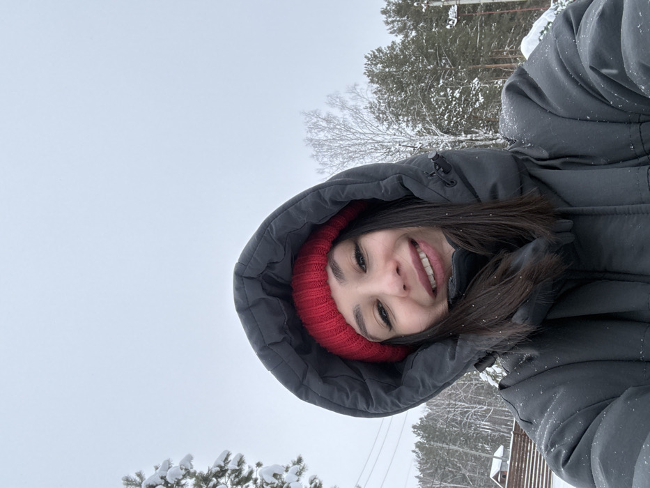 A female subject is centered in a close-up, upward-angled photograph, consistent with a selfie, taken outdoors in Sysert, Russia. She wears a red knitted beanie and a dark grey or black hooded puffer jacket, with the hood pulled up around her head. Her dark hair is visible beneath the beanie. Her face is presented with a smile, revealing teeth. Sparse white particles, consistent with snowflakes, are visible on her jacket and hair. The background depicts a snow-covered winter environment under an overcast, light grey sky. To the subject's left, a snow-covered coniferous tree is partially visible, along with power lines. Further back and to the left, a portion of a house with a light-colored exterior and a dark, snow-covered roof is seen behind a reddish-brown wooden fence. The right side of the background features a snow-laden landscape with both coniferous and bare deciduous trees, and a utility pole with lines. The ground throughout the visible background is covered in snow.
