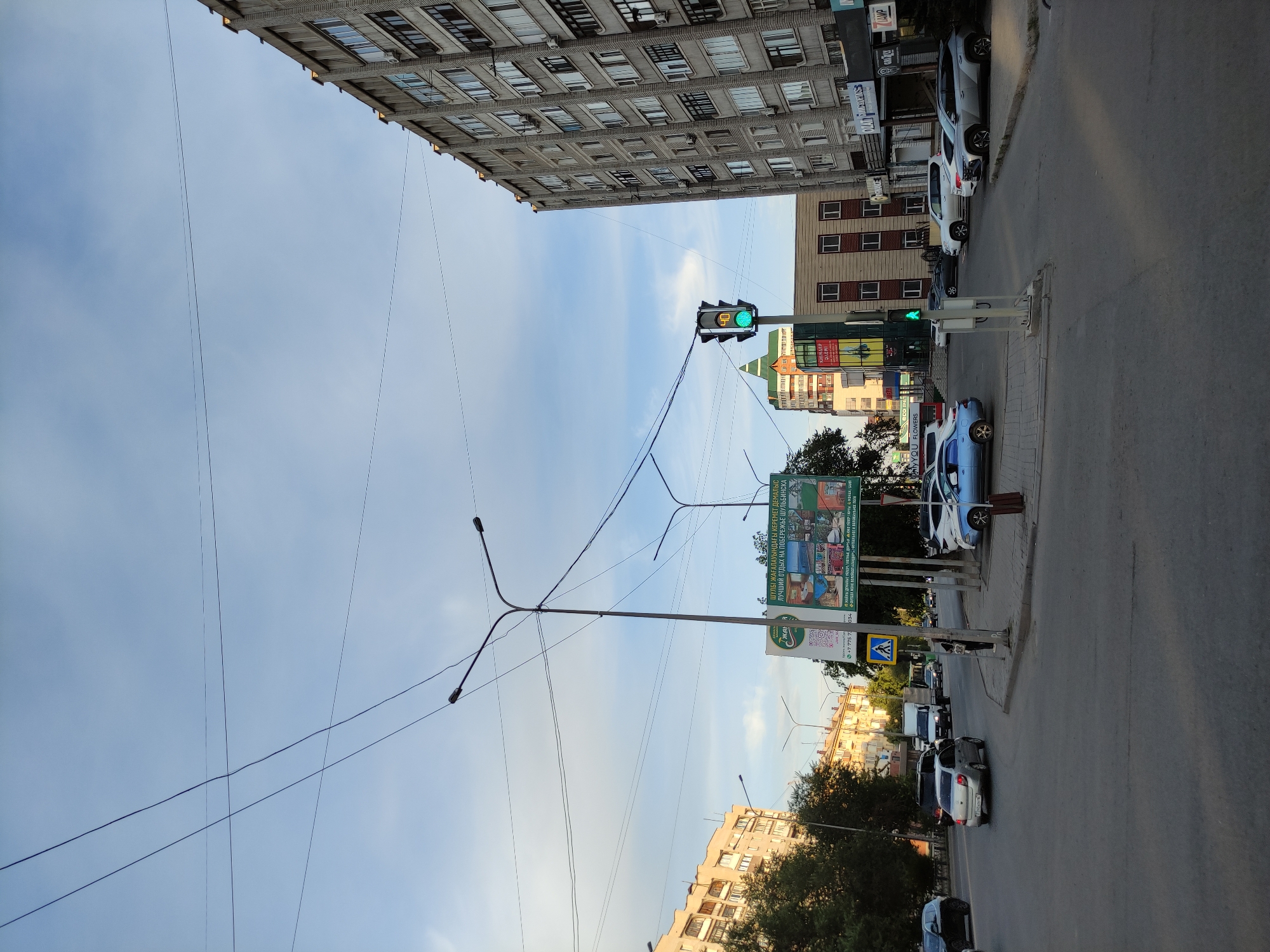 The image depicts a street intersection in the city of Semey, Kazakhstan, on a clear day with a partly cloudy sky. The perspective is from a street level, looking towards a multi-lane intersection. Several buildings line the street, including a tall, modern apartment building on the right and a lighter-colored, multi-story building on the left. A billboard advertising a holiday resort is visible in the mid-ground. Traffic lights are operational, with a green light illuminated on one. Vehicles, including cars and a truck, are present on the road, suggesting moderate traffic. Power lines crisscross the sky above the intersection, supported by street lamps. The overall scene suggests a typical urban environment in Semey.