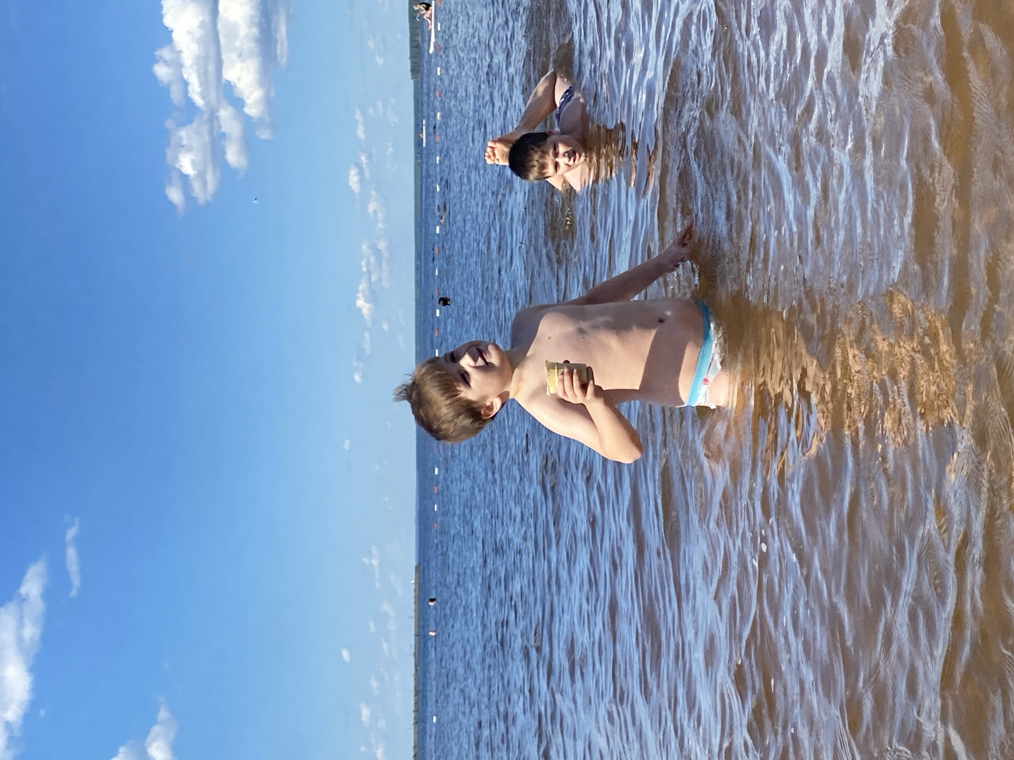 The image depicts two young boys in the water on a sunny day, likely at a beach or lake. The boy in the foreground, on the left, is partially submerged, holding a light-colored object, possibly an ice cream cone, in his right hand. He is looking upwards and to his right. He is wearing blue and white patterned swim trunks. The second boy is further back in the water, lying on his back with his legs in the air. He is looking towards the viewer. The water is blue and appears calm with some ripples. In the background, there is a long stretch of water with a distant shoreline. There are buoys marking a swimming area. The sky is blue with scattered white clouds. The scene suggests a recreational setting, possibly a public beach. The location is identified as the city of Chaikovsky, Russia.