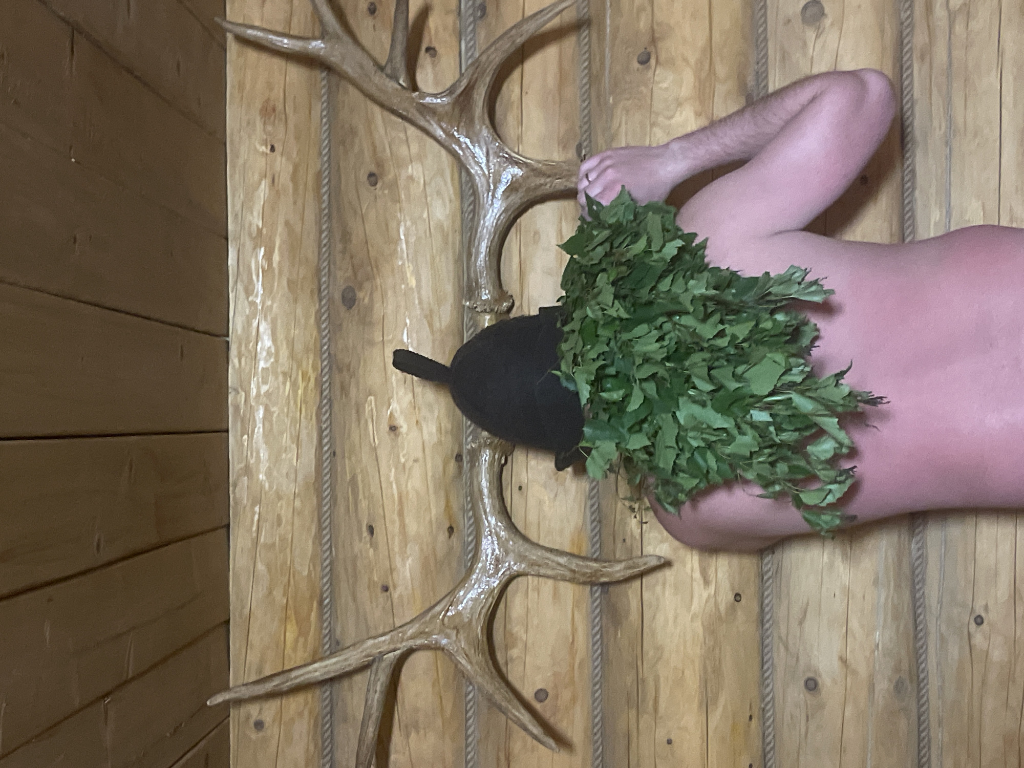 A person lies on their back on a light-colored wooden surface with rope details, potentially a bench or floor in a sauna or similar setting. The person wears a black sauna hat with a tassel and a bundle of green leafy branches partially covers their chest. Above them are two sets of deer antlers mounted on the wall. The location is likely Kuyeda, Russia, based on the image context.