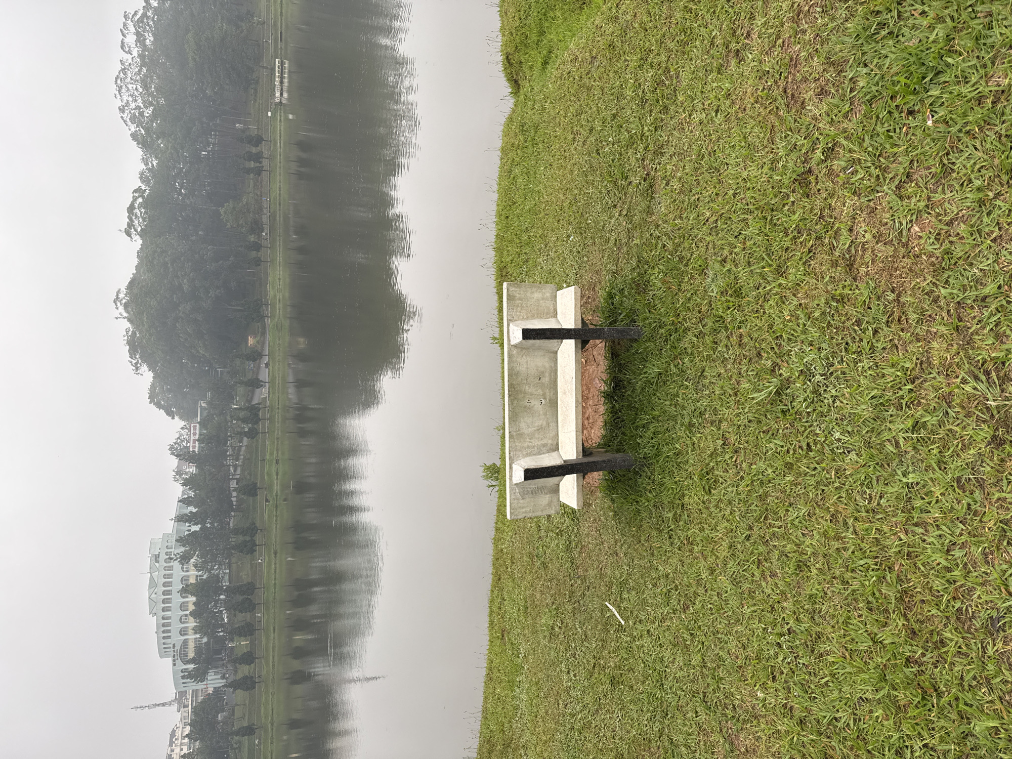 The image captures a peaceful lakeside scene in Da Lat, Vietnam, under an overcast sky. The foreground features a simple concrete bench with black armrests, positioned on a vibrant green grassy bank next to the water's edge. The grass shows some drier patches. The expansive body of water, likely a lake, is calm and acts as a mirror, reflecting the hazy outlines of the distant shore.
On the opposite side, a dense line of tall green trees stretches along the bank, and further along, a large, light-colored building with distinctive arched windows and multiple stories is partially visible, also reflected in the water. The overall impression is one of tranquility and quiet, with the muted light suggesting either early morning or a heavily overcast day, possibly with mist or fog in the distance. No people are present, and there is no discernible text in the scene.