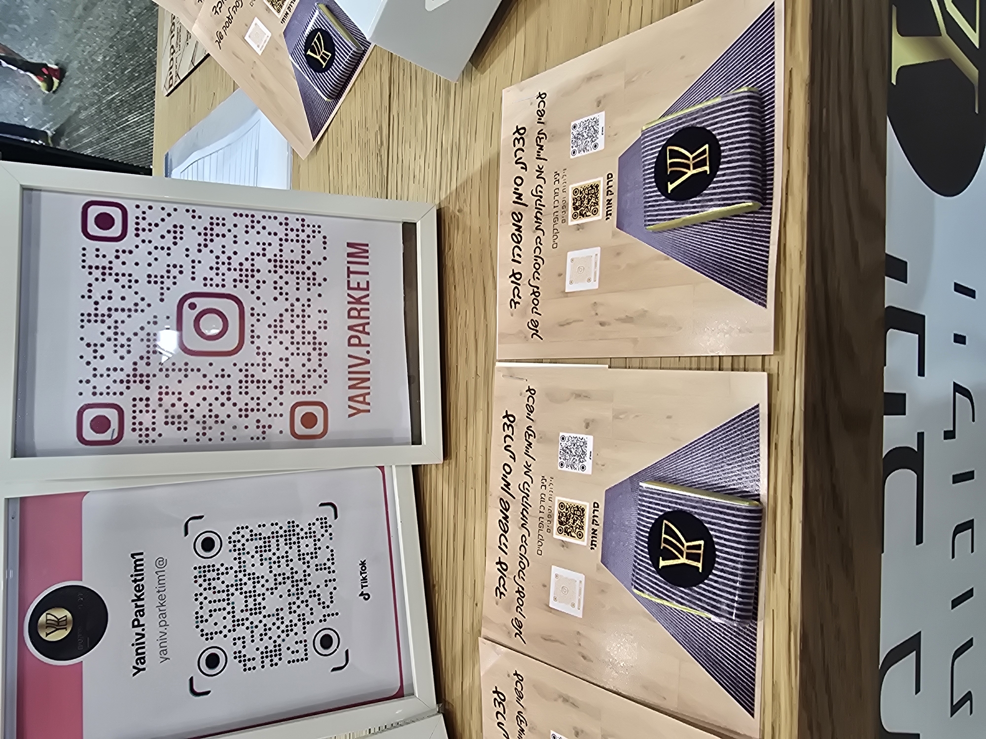 The image shows two framed signs and multiple identical printed cards arranged on a wooden surface. The top framed sign displays an Instagram QR code and the text "YANIV.PARKETIM". The bottom framed sign features a TikTok QR code and the text "Yaniv.Parketim1 yaniv.parketim1@". Each printed card has a design incorporating a logo with a stylized "Y", a QR code, and Hebrew text indicating "Yaniv – The Center of Parquet Flooring, Curtains, and Wallpaper". The text also invites customers to "Come get a recommendation from the flooring experts".
The image suggests that the scene is related to a business promotion or advertising for "Yaniv Parketim", a company that deals with flooring, curtains, and wallpaper, possibly based in Tel Aviv, Israel, given the Hebrew text on the cards.