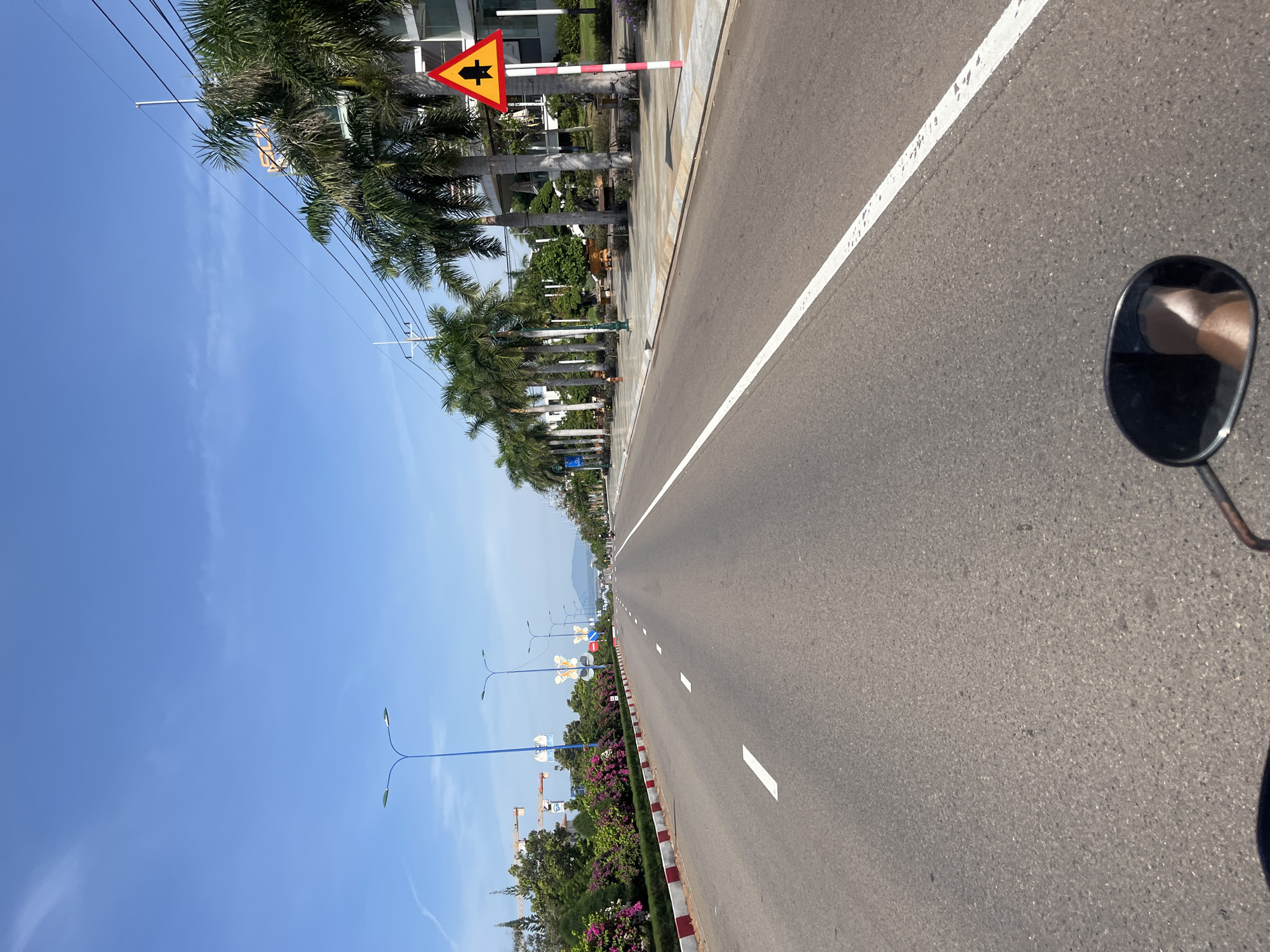 The image shows a wide asphalt road with white lane markings, stretching into the distance under a clear blue sky. The road is lined with palm trees on the right and a landscaped area with flowering bushes and more trees on the left. Modern buildings and streetlights are visible along the sides. A cautionary traffic sign, a yellow triangle with a black symbol indicating a junction or intersection, is posted on the right. In the foreground, a portion of a motorcycle's side mirror, reflecting a person's leg, is visible. The scene appears to be in a well-maintained urban or suburban area, possibly a coastal road, given the palm trees. The general atmosphere is bright and sunny. The location is identified as Phu Thuy Ward, Vietnam.