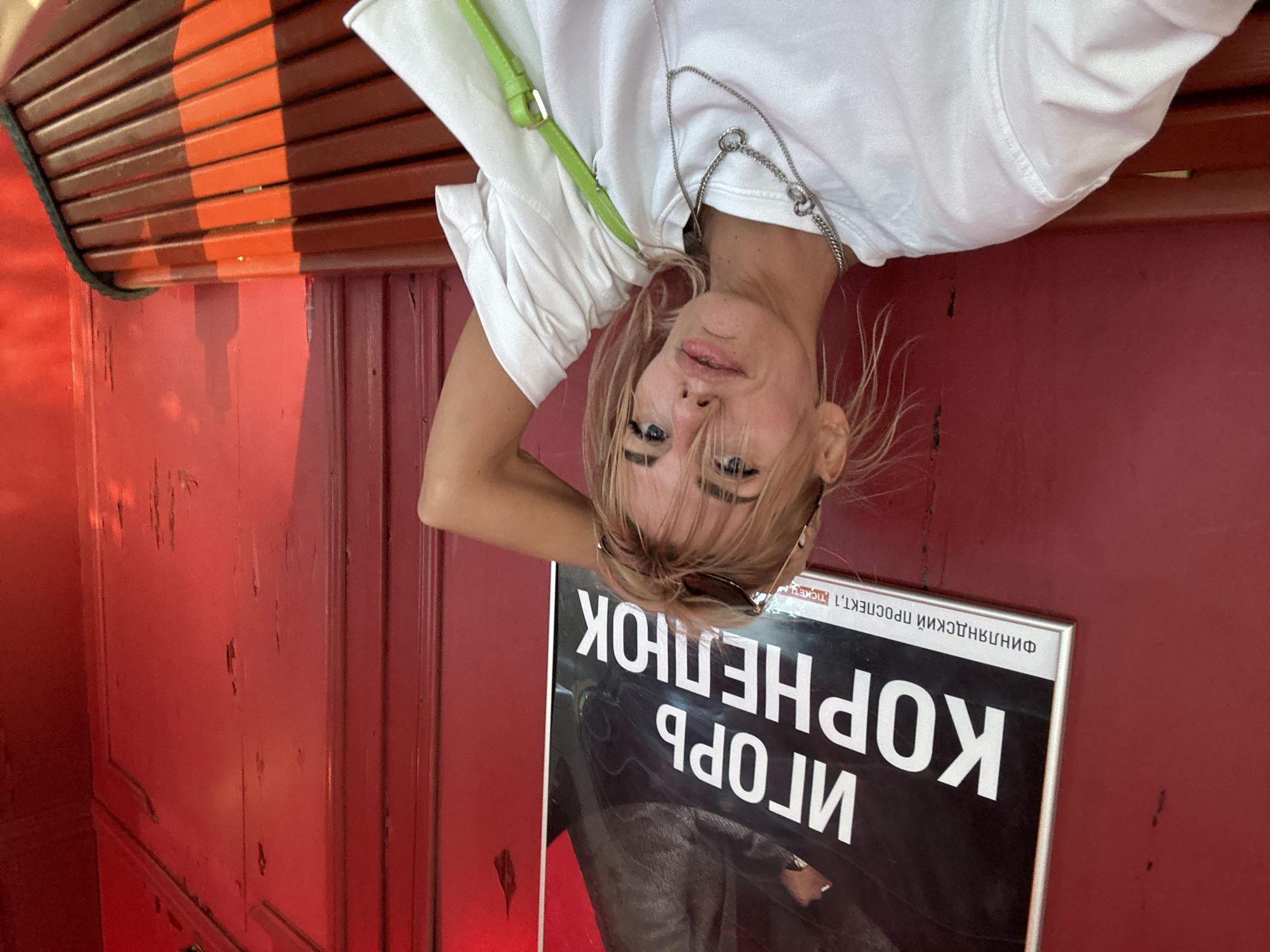 A young Caucasian woman with blonde hair is pictured in a medium shot, seated on a wooden bench with a red background. She is wearing a white t-shirt and a green purse is visible. She has a silver necklace on and sunglasses pushed up on her head. Her right arm is raised behind her head. Behind her, a framed poster with Russian text, which appears to be an advertisement, is partially visible. The setting seems to be an outdoor area, possibly a waiting area or a public space, given the bench and the poster. The red background suggests it might be a building or a structure painted red. The lighting indicates it is daytime.