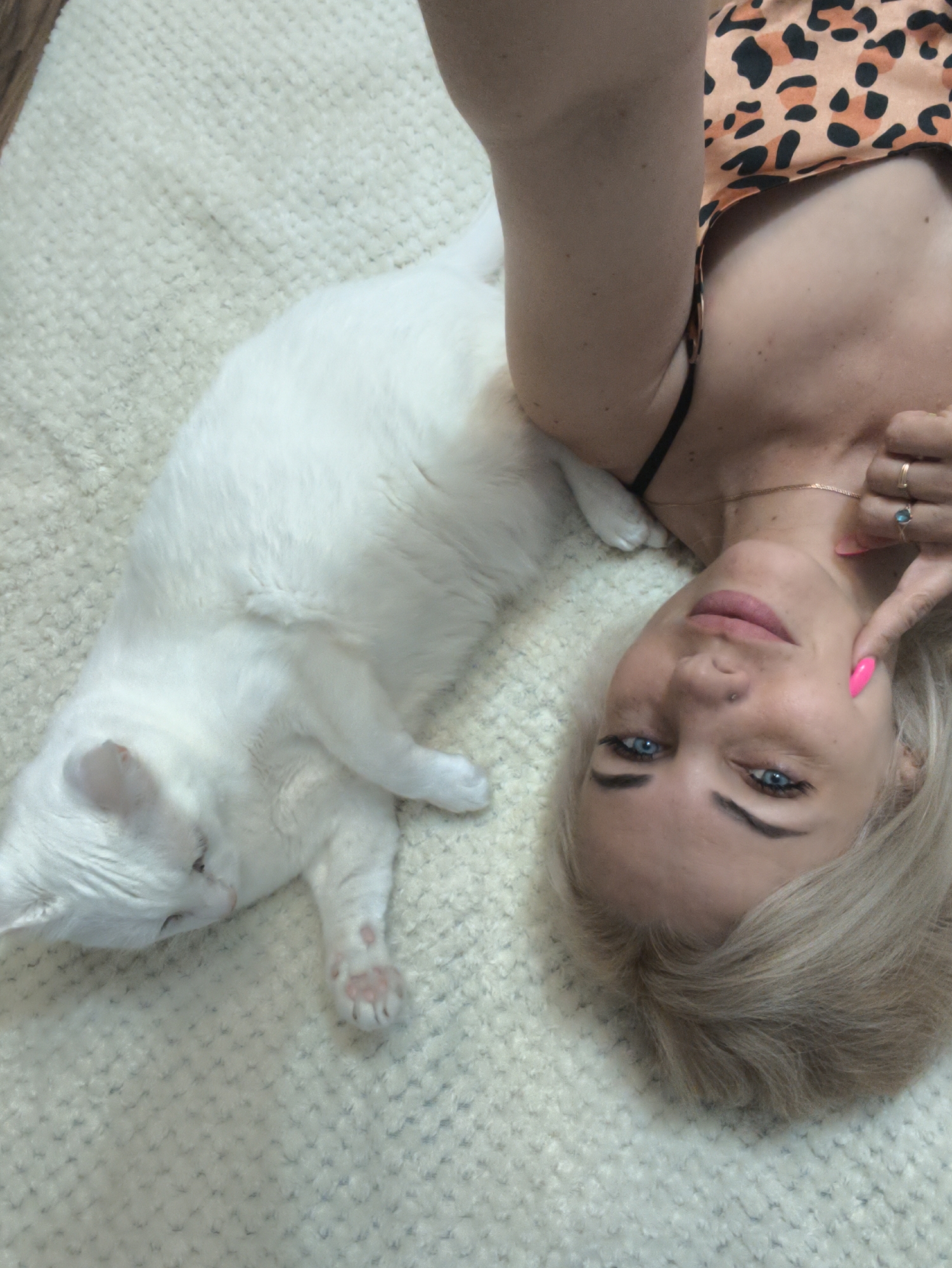 A blonde woman with blue eyes lies on a white textured surface next to a white cat. The woman wears a leopard-print top and has pink nails. She touches her cheek with a finger. A gold necklace is visible on her neck. The cat lies on its side, its paws extended. The background is the white textured surface. The location is not discernible from the image. The city of Sevastopol, Ukraine is not discernible from the image.