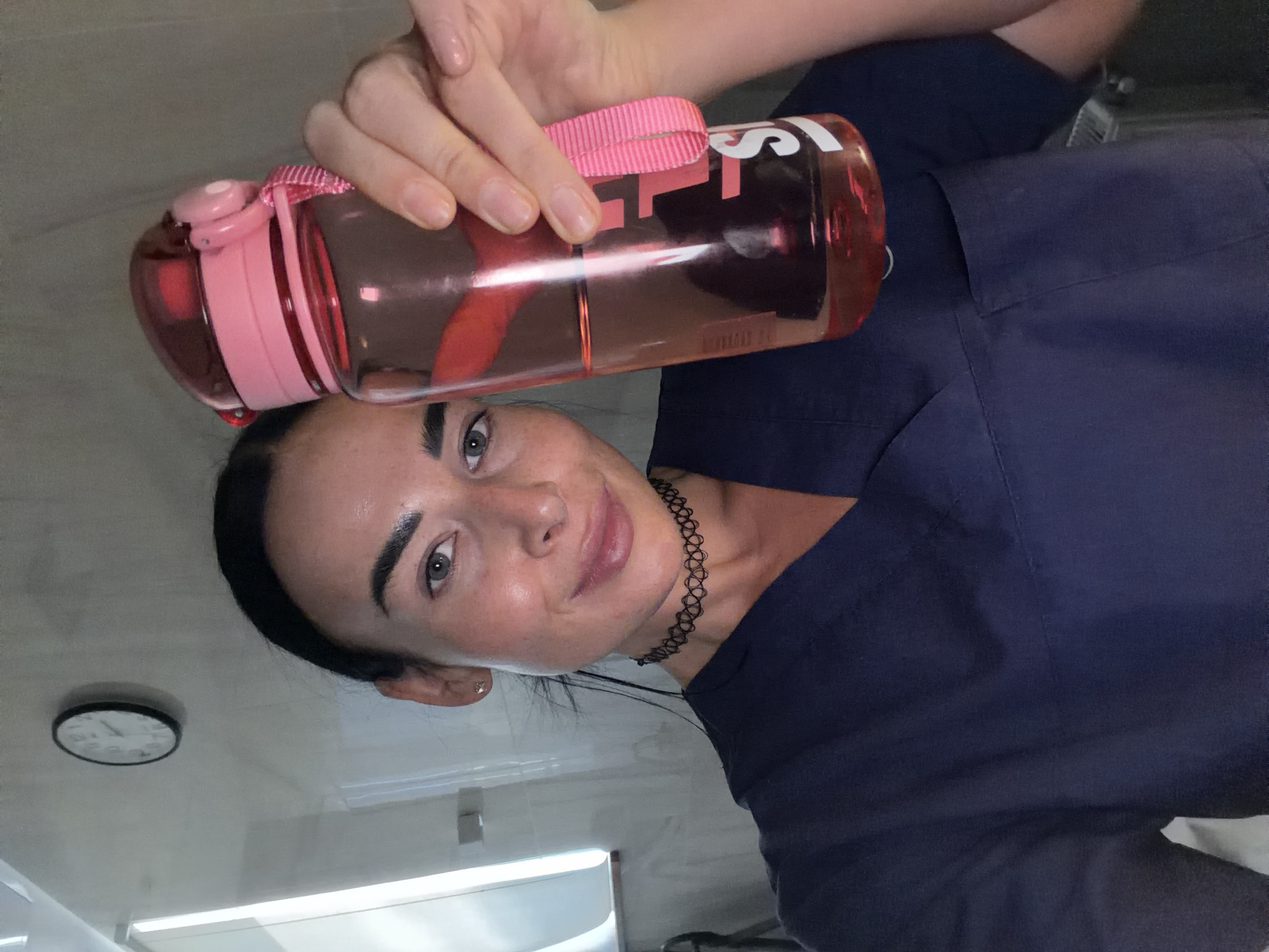 This portrait-style image, originally rotated 90 degrees counter-clockwise, depicts a young woman taking a selfie. She has dark hair pulled back, prominent eyebrows, and light-colored eyes, looking towards the camera with a subtle, pleasant smile. She is wearing a dark blue top, which appears to be a uniform or scrubs, and a black choker necklace.
She is holding a clear water bottle with pink plastic accents on the lid and a pink fabric strap. The bottle contains a pinkish-red liquid. Partially visible white text on the bottle's side shows letters such as "S" and "IST".
The background indicates an indoor setting with plain white walls and artificial overhead lighting. A round, analog wall clock is visible to the woman's left, displaying a time that appears to be around 11:30 or 12:30. The environment suggests a functional space, such as a clinic, office, or break room. The woman's pose, holding the water bottle, implies a moment of hydration or a short break. The location context provided for the image is Saint Petersburg, Russia, though no specific visual cues within the image explicitly confirm this.