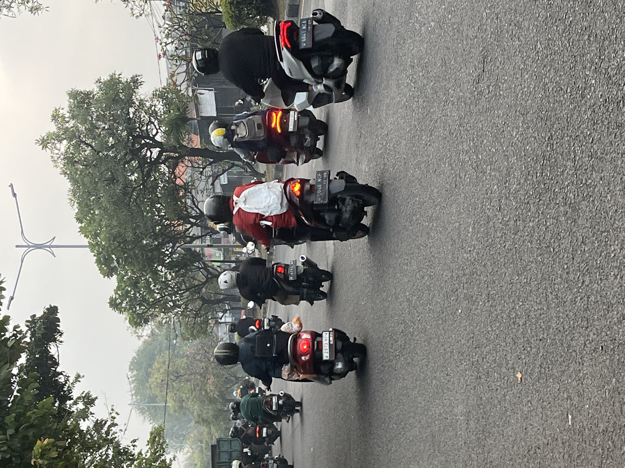 A group of people are riding motorcycles on a paved road in Sumorame, Indonesia. The sky is overcast, and the scene appears to be in the morning or late afternoon. The motorcyclists are wearing helmets and protective gear. Some are carrying backpacks or other items. The motorcycles are of various makes and models. Trees and buildings are visible in the background. The road is wet, suggesting it has recently rained.