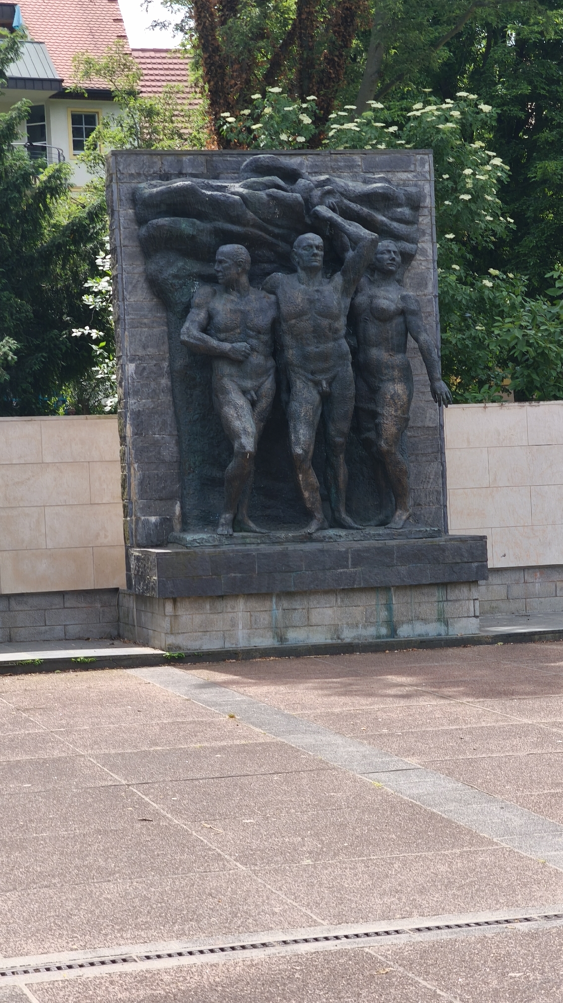 The image shows a bronze relief sculpture on a stone monument. The sculpture depicts three nude male figures in what appears to be a struggle or a shared journey. The figures are muscular and are set against a textured, abstract background that could represent waves or clouds. The monument itself is made of light-colored, rectangular stone blocks and a darker stone base. The sculpture is positioned on a paved area composed of reddish-brown, textured square tiles. The background includes greenery and partial views of buildings with a red roof, indicating an urban or park setting. The location is an Unknown city.