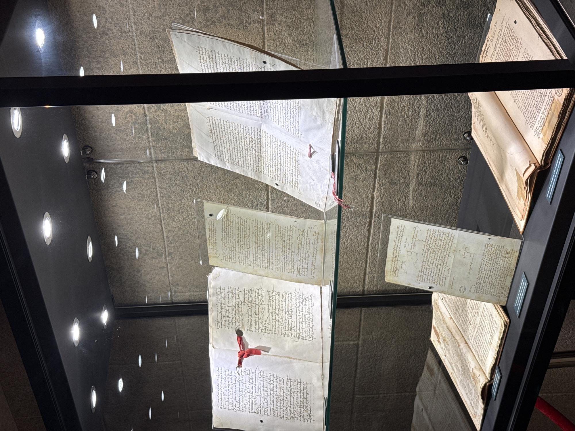 In this media file, ancient manuscripts are displayed within a glass-enclosed exhibit. The scene is lit by overhead lights, creating bright circular reflections on the dark interior surface of the display case, which also reflects the textured stone wall behind the exhibit.
The primary objects are several unfolded manuscripts, appearing as aged parchment covered in dense, handwritten text in a script that is likely Arabic or a related language. One manuscript, positioned at the top of the frame, is spread open and held by clear acrylic panels. Below it, another manuscript is similarly displayed, with a vibrant red ribbon tied around it, suggesting it is a significant artifact. Further down, more pages of text are visible, some appearing as parts of a larger book or scroll. To the right, an open book with yellowed pages and similar script is also on display.
The overall impression is one of historical preservation and academic display. The lighting and reflective surfaces suggest an indoor museum or archive setting. No people are visible, emphasizing the focus on the historical documents. The image captures the quiet reverence of viewing ancient texts, with the surrounding environment designed to protect and showcase these fragile pieces of history. The specific location, Benalmádena, Spain, is mentioned, but no visual cues within the image directly confirm this location beyond it being a display within a Spanish context.
