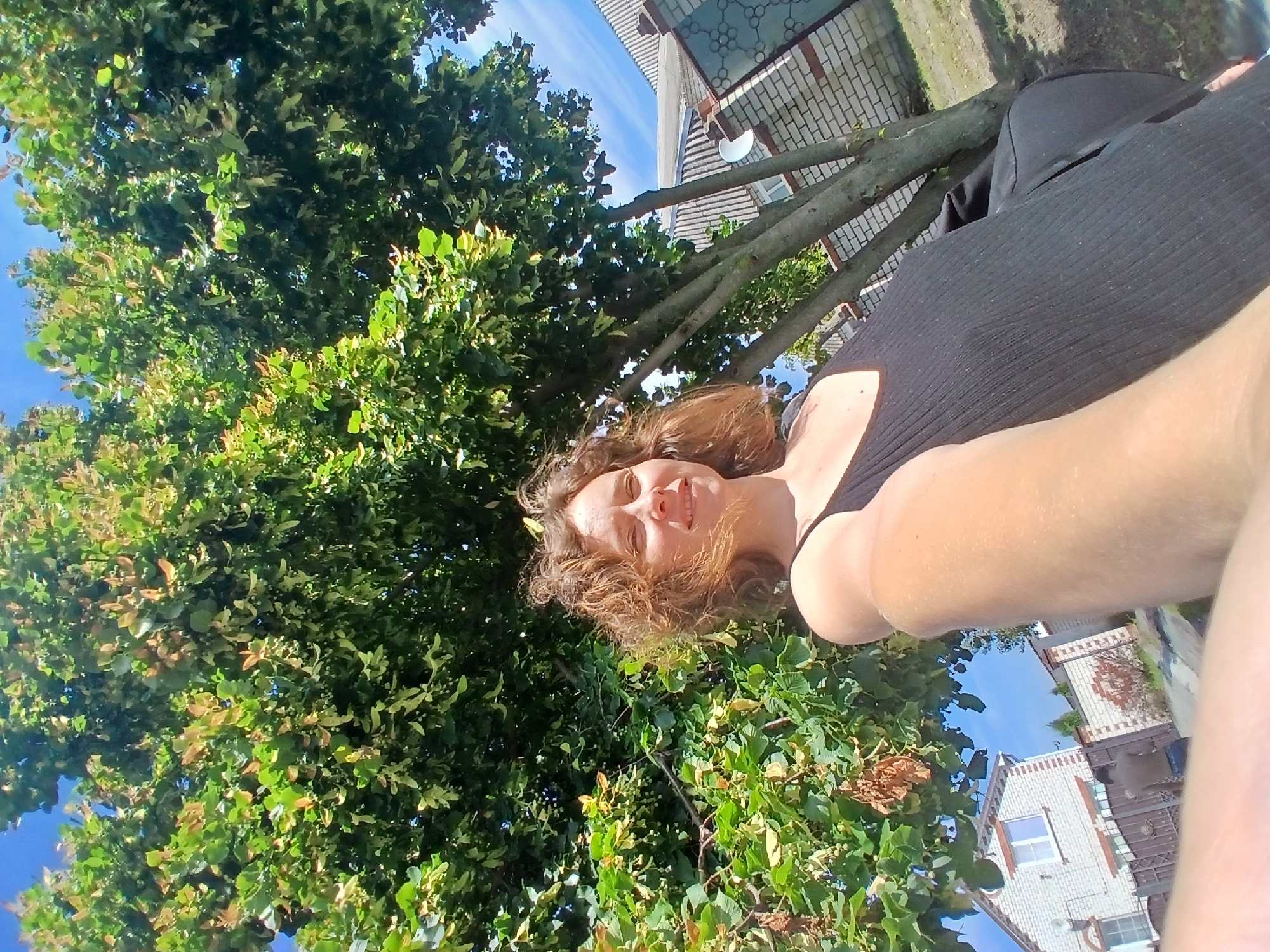 A woman with curly brown hair is taking a selfie outdoors under a large tree with green leaves. She is wearing a black tank top and is smiling at the camera. In the background, there is a brick fence, a house, and a bright blue sky with some clouds. The scene appears to be in a residential area.